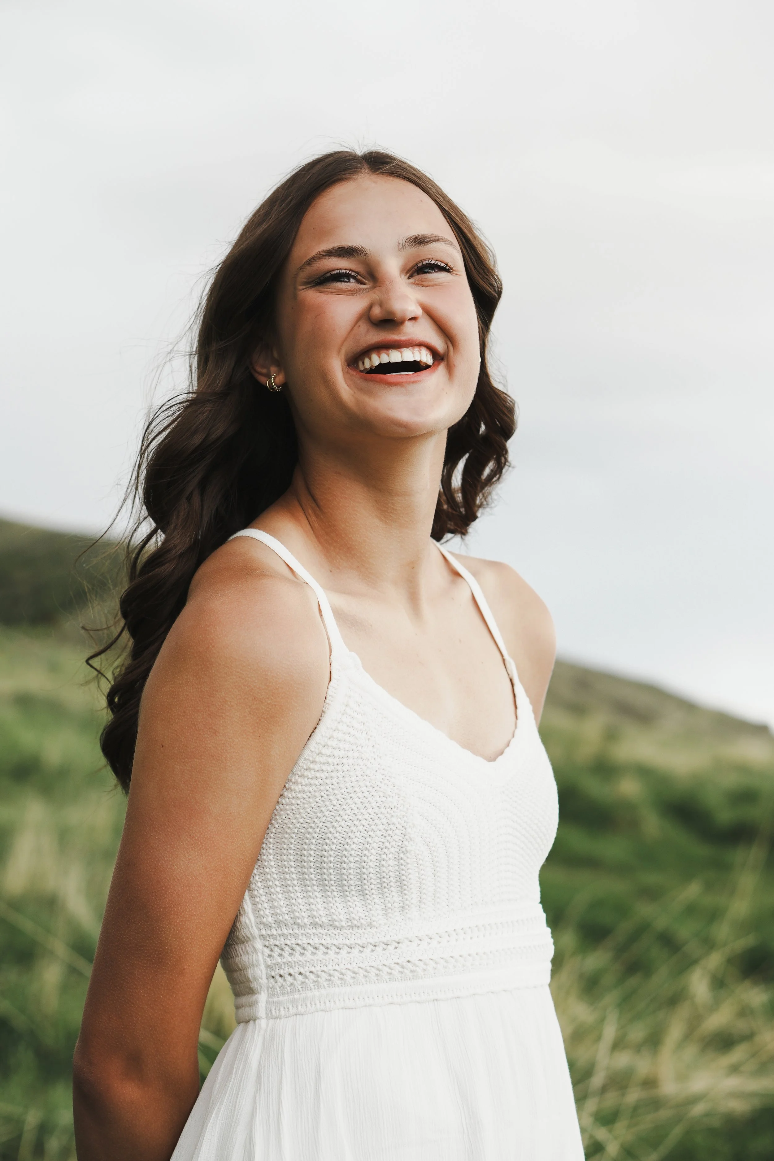 A young woman with long dark hair, wearing a white sundress with spaghetti straps, standing outdoors in a grassy area with a hill in the background, smiling and looking happy.