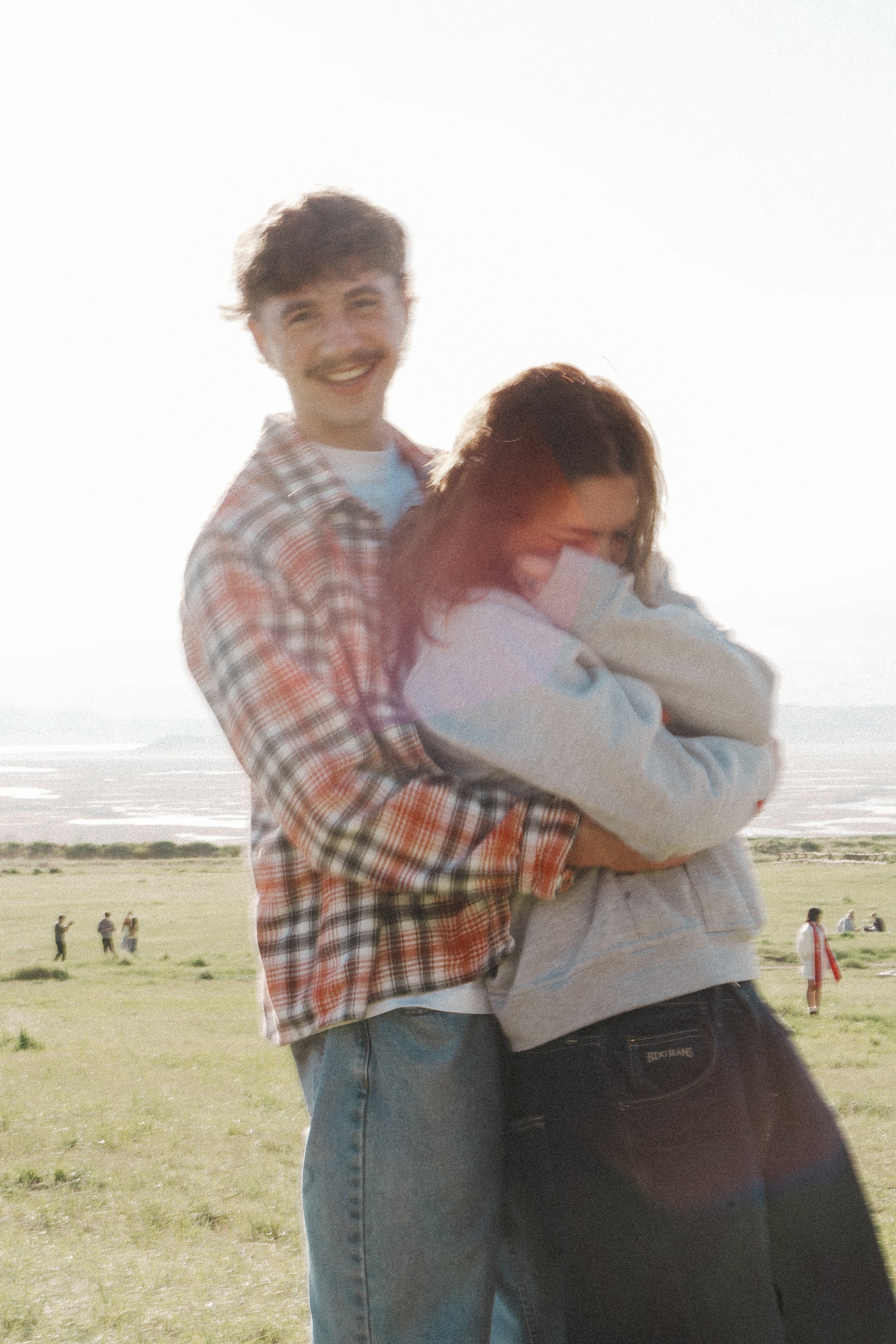 A young man and woman embrace outdoors in a grassy field, with a body of water and a bright sky in the background. The man is smiling, wearing a plaid shirt and jeans, and the woman has her face turned away, wearing a sweatshirt.