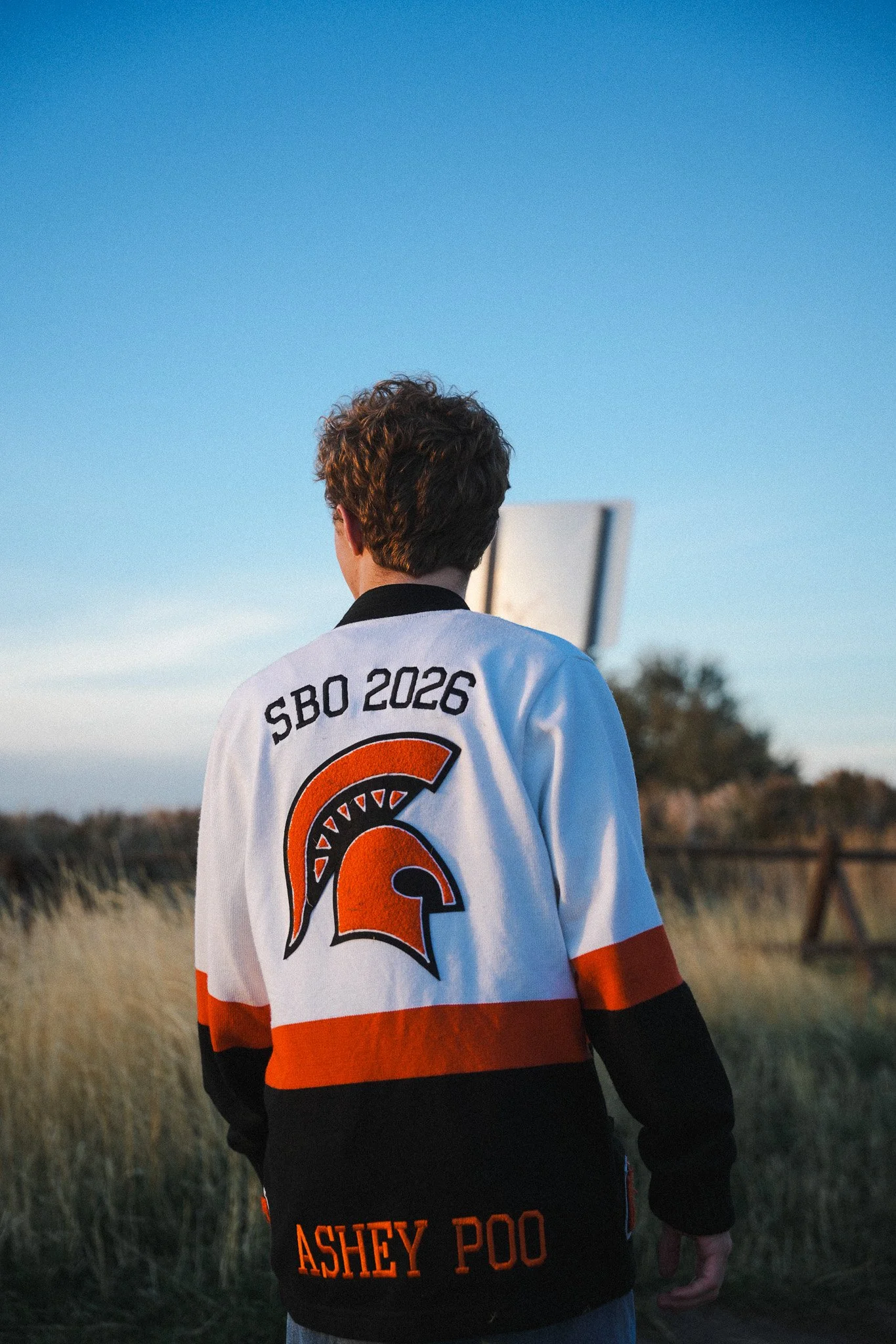 Person wearing a white, orange, and black jacket with a Spartan helmet logo and 'SBO 2026' and 'Ashley Poo' text, standing outdoors on a grassy field with a blue sky and distant trees.