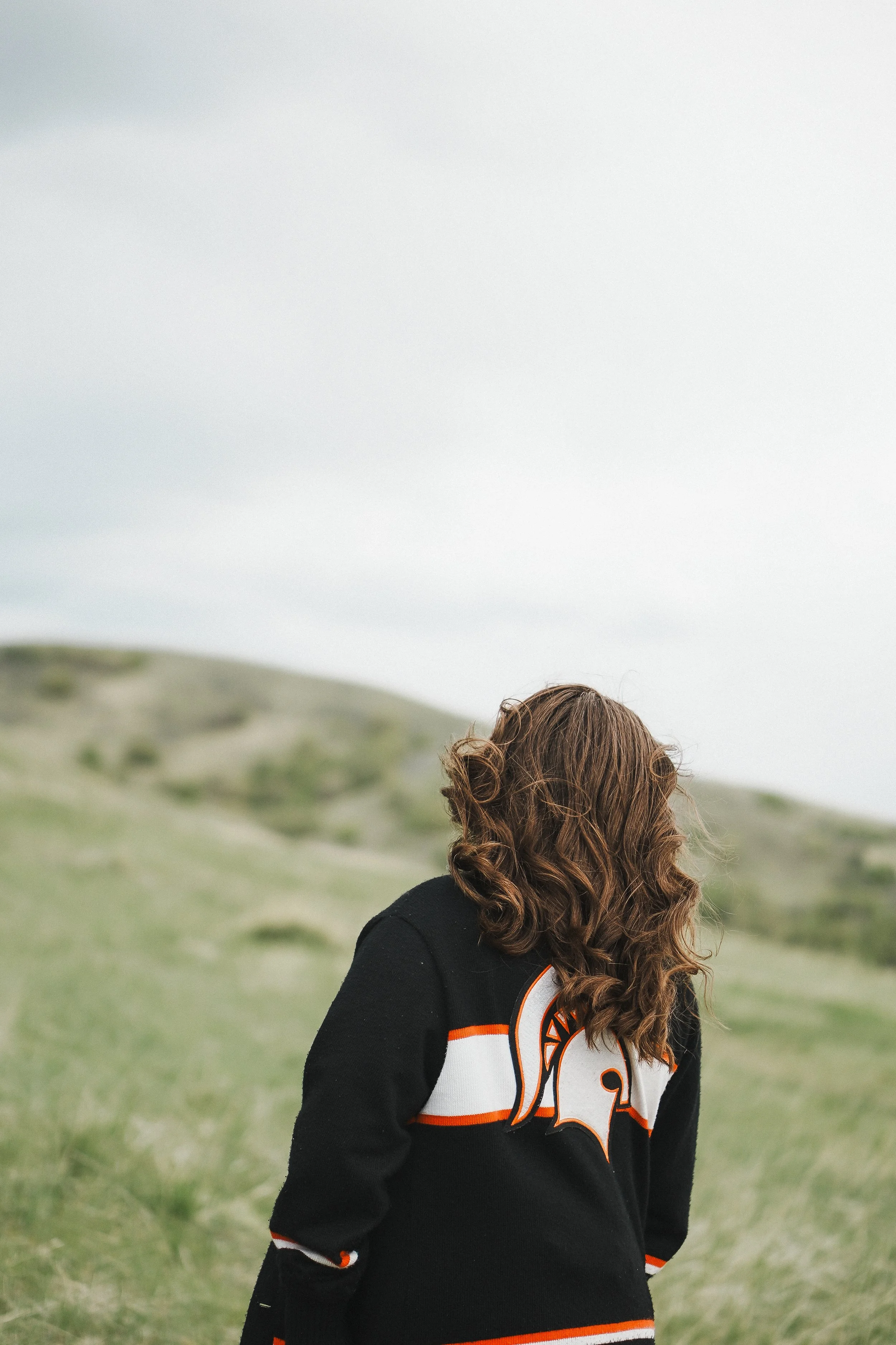 A woman with brown curly hair standing outdoors on a grassy hill under a cloudy sky, wearing a black and white sports jacket with orange accents and a logo.