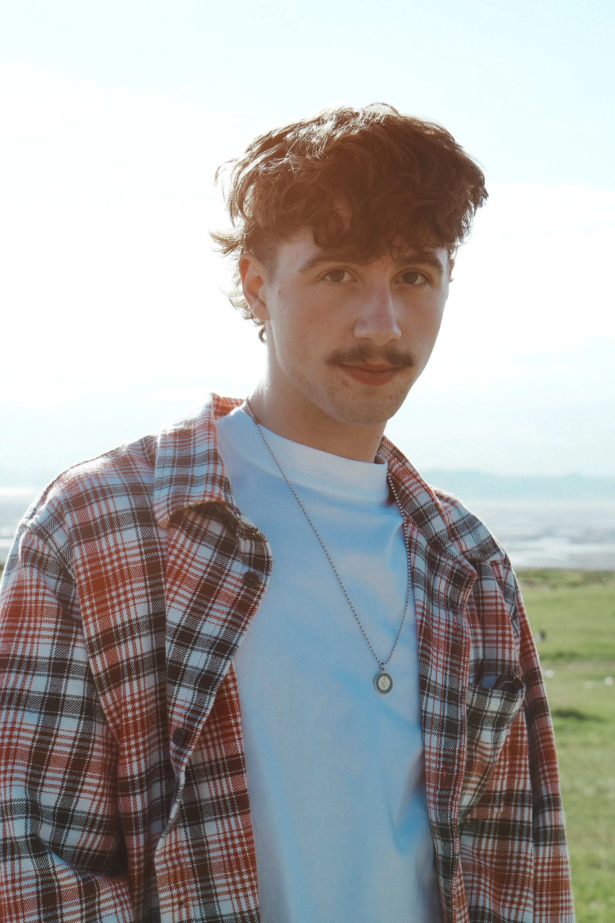 A young man with curly hair, a mustache, and light skin standing outdoors on a sunny day near the coast, wearing a white t-shirt, a plaid shirt, and a necklace with a circular pendant.