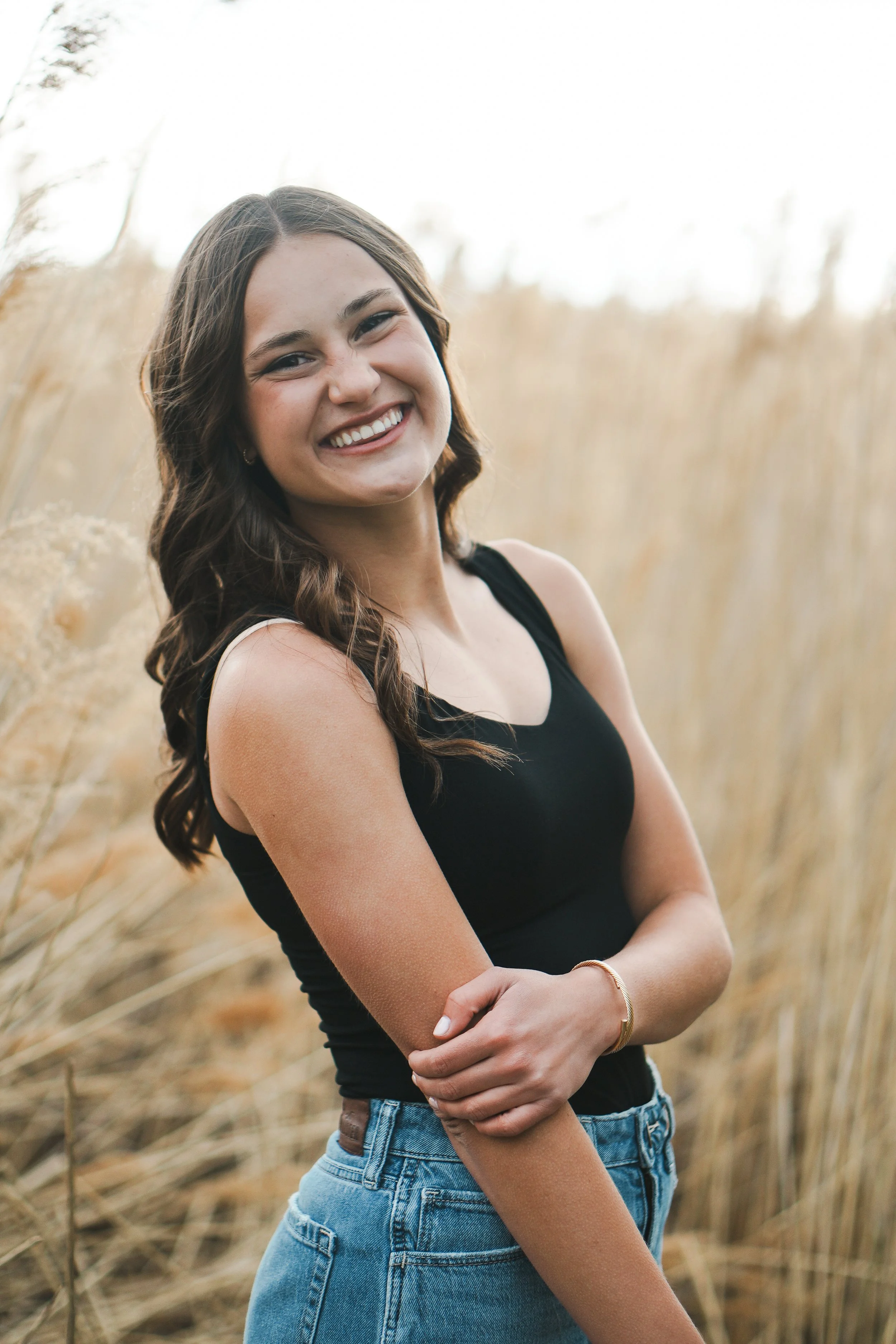 A young woman with brown hair smiling outdoors in a field of tall, golden grass.