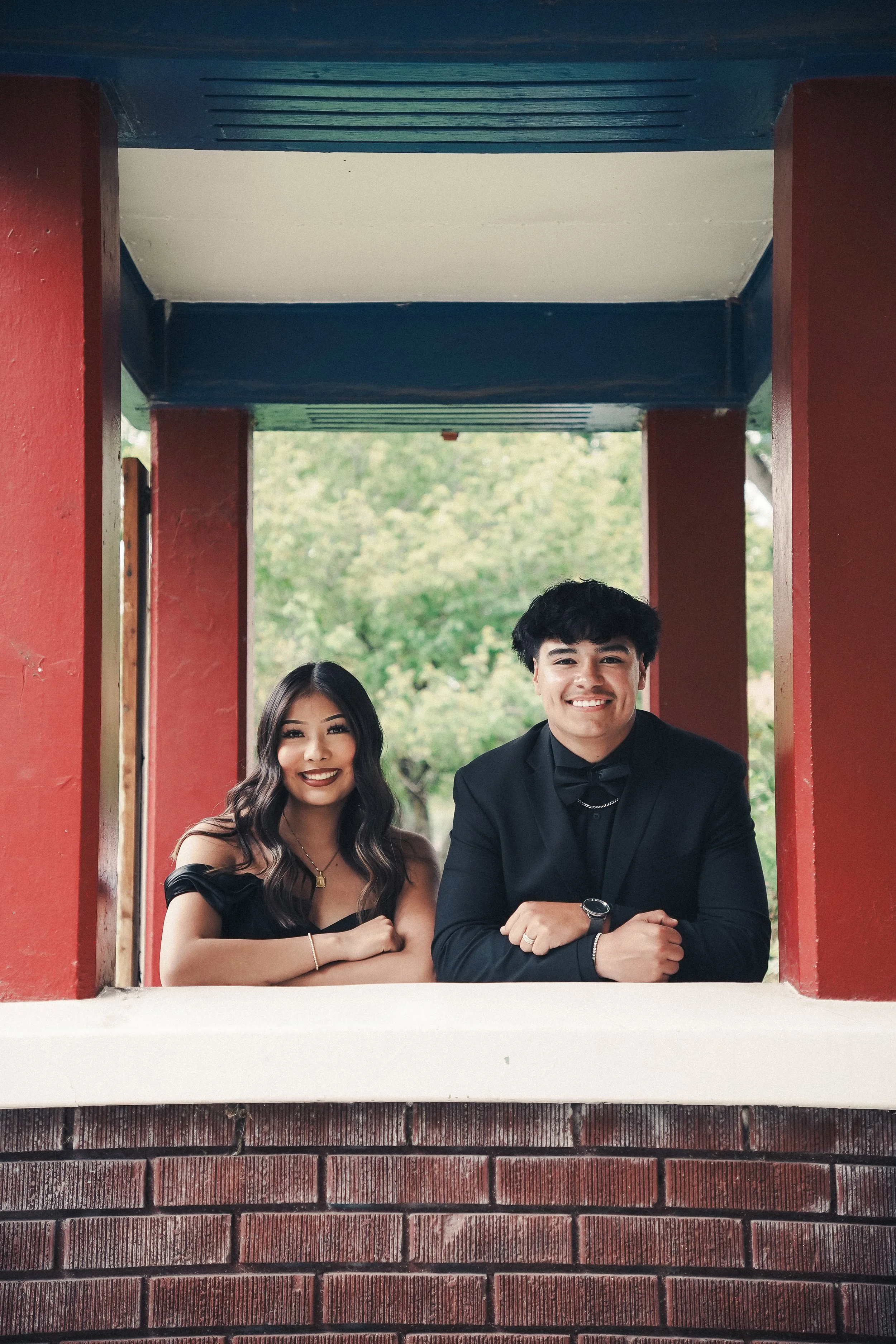 A young woman and a young man in formal attire smiling and posing behind a brick railing, with a green outdoor background.