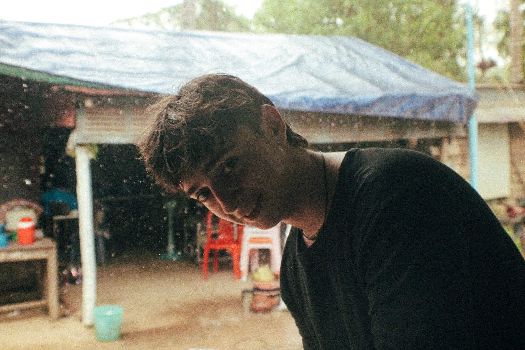 A young man with dark hair smiling and looking at the camera, leaning to one side under an outdoor shelter with a blue tarp in the background, with various objects and chairs nearby.