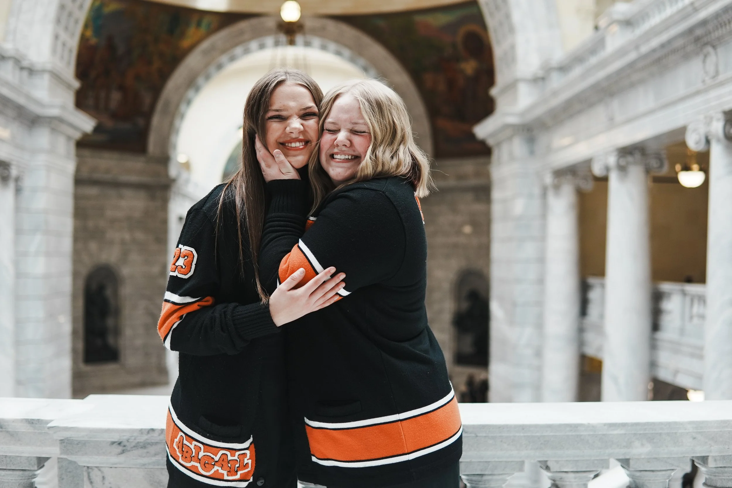 Two young women hugging and smiling in a grand, ornate building with arches and columns.
