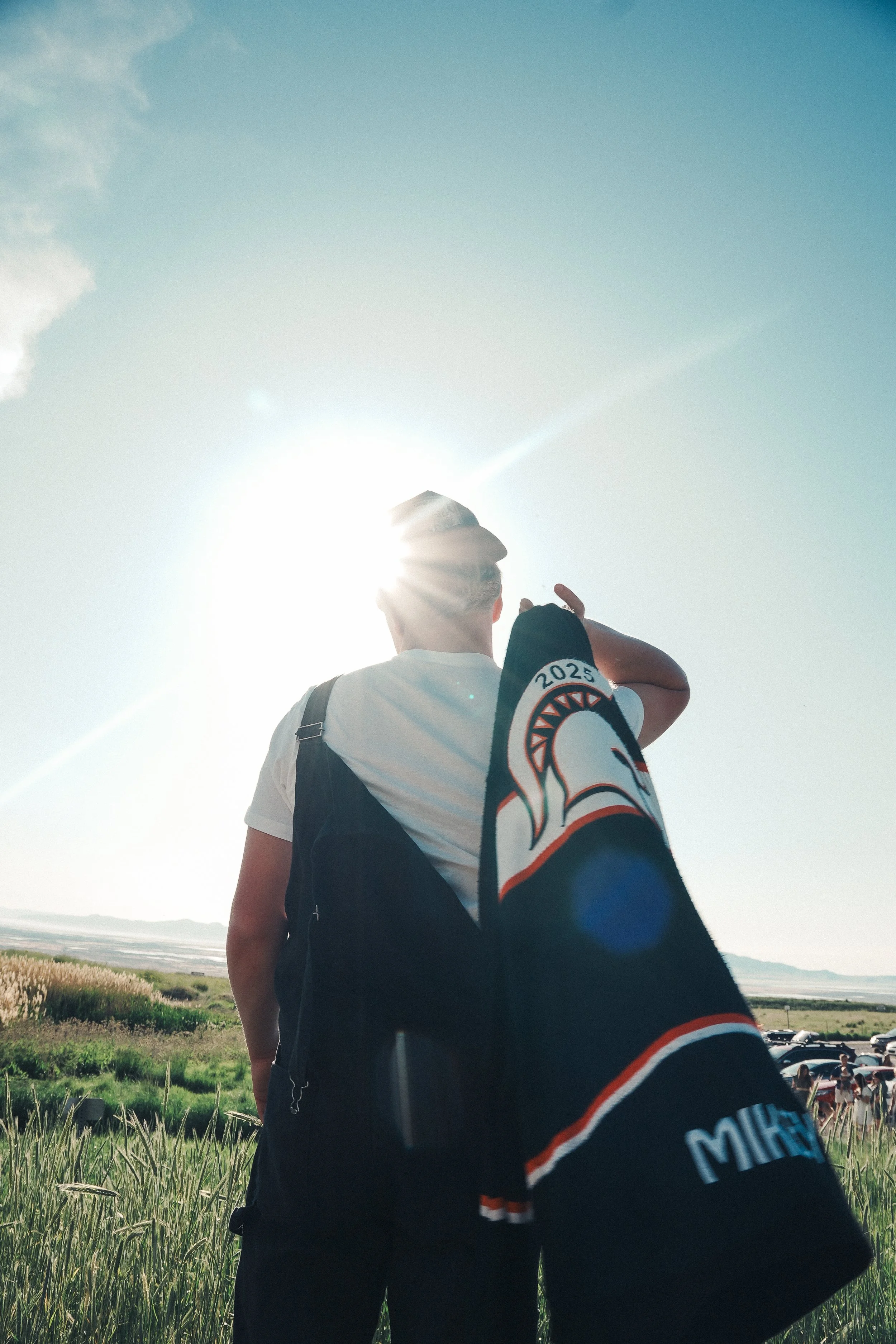 A person wearing a white t-shirt and a baseball cap stands outdoors in a grassy field, carrying a black backpack and a black jacket with a logo and the year 2025, gazing towards the bright sun in a clear sky.