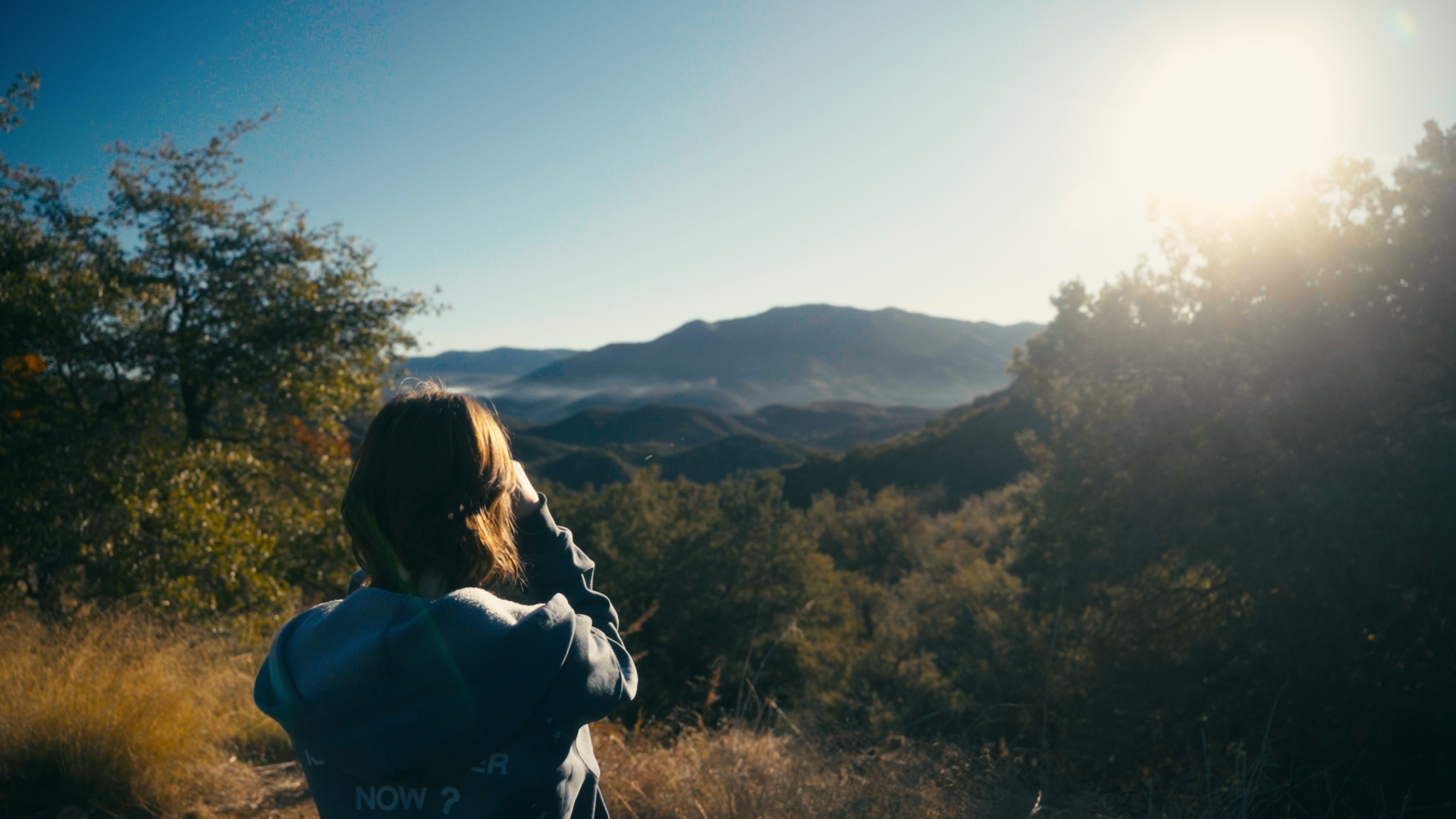 A person standing outdoors on a hillside, looking at a landscape of mountains and trees during sunset.