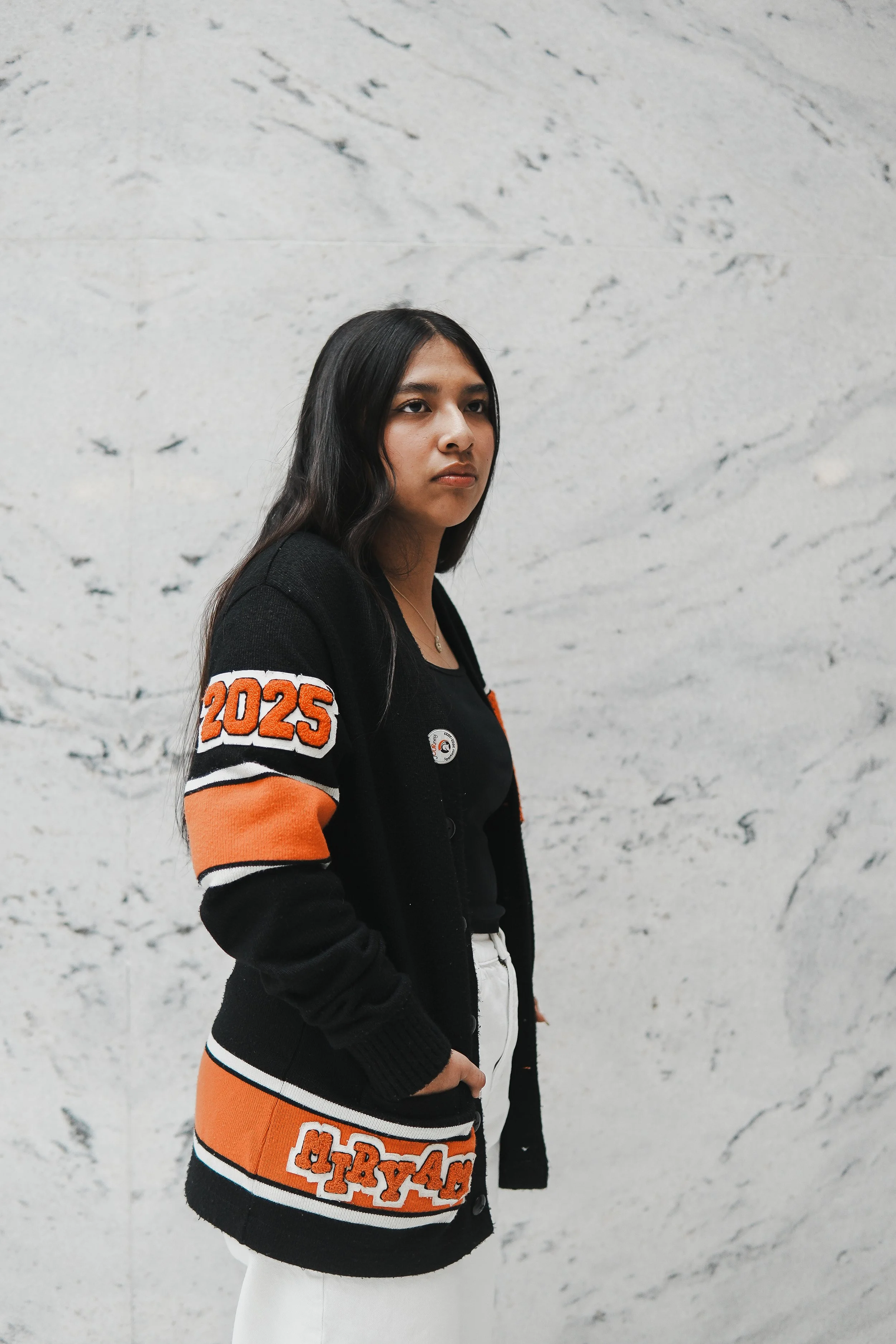Young woman with long dark hair standing against a light gray marble wall, wearing a black and orange varsity-style sweater with the year 2025 and other patches, and a black top underneath.