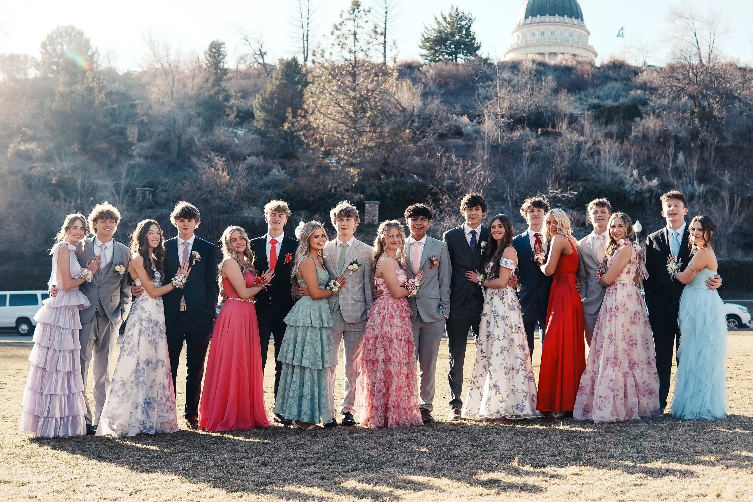 Group of young people dressed in formal attire, standing outdoors on grass, celebrating a special occasion with a building and trees in the background.