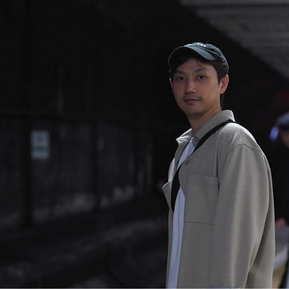 Young man with a beige jacket, white shirt, and black cap standing in a dark, possibly underground, tunnel or subway area.
