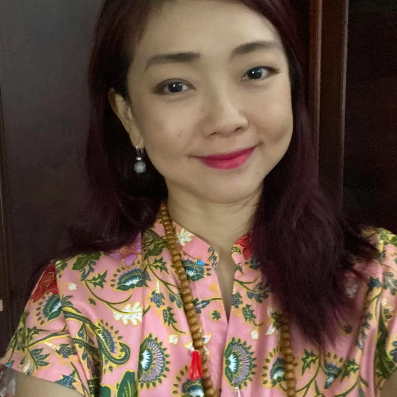 A woman with shoulder-length dark red hair, wearing earrings, a beaded necklace with a red tassel, and a pink floral-patterned top, smiling in front of a dark wooden background.