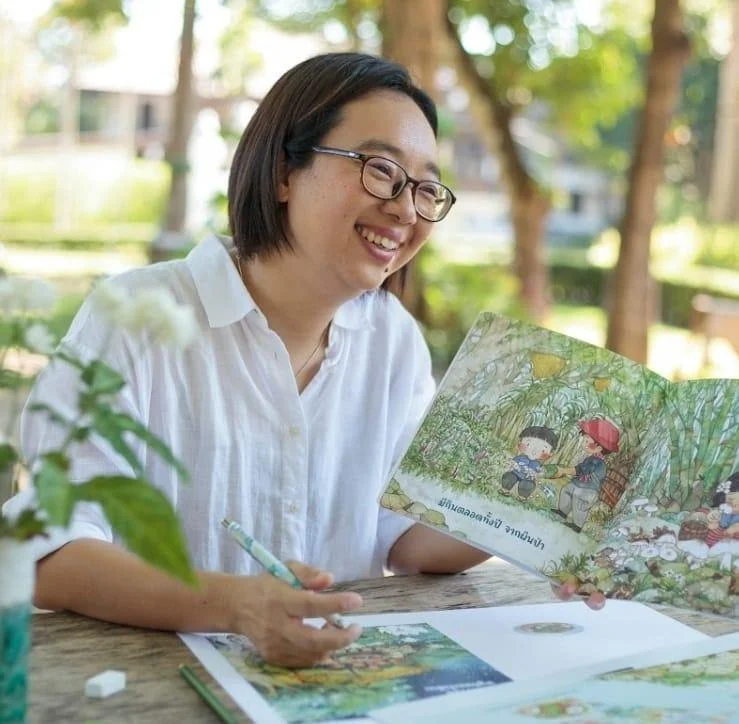 A woman with glasses and a white shirt smiling while reading a colorful children's book outdoors at a wooden table.