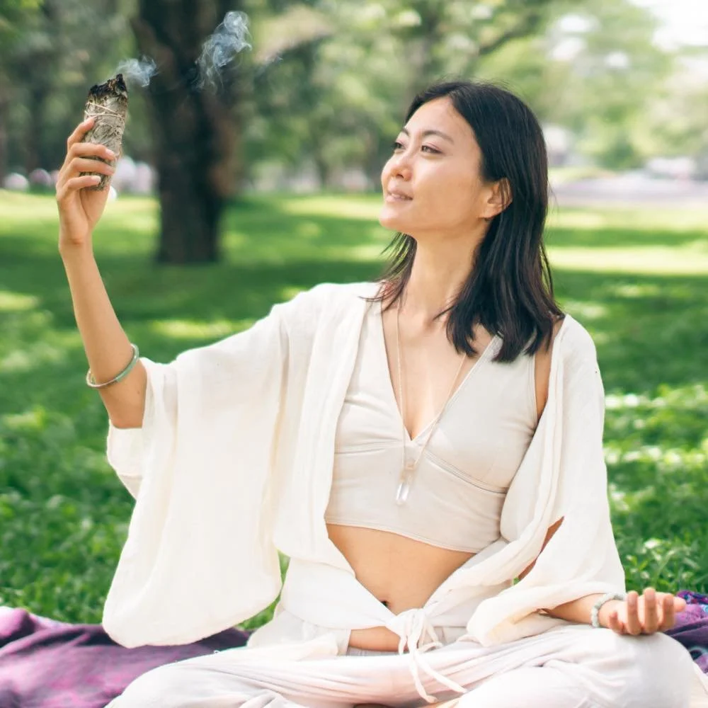A woman practicing meditation outdoors in a park with green grass and trees, holding a burning sage bundle in her right hand and sitting on a blanket.