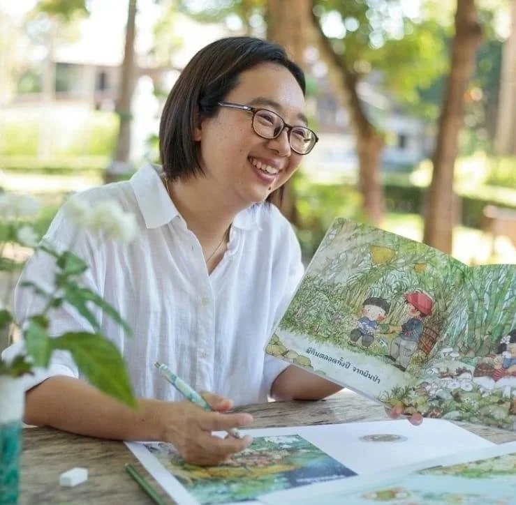 A woman with glasses and a white shirt smiling while reading a colorful children's book outdoors at a wooden table.