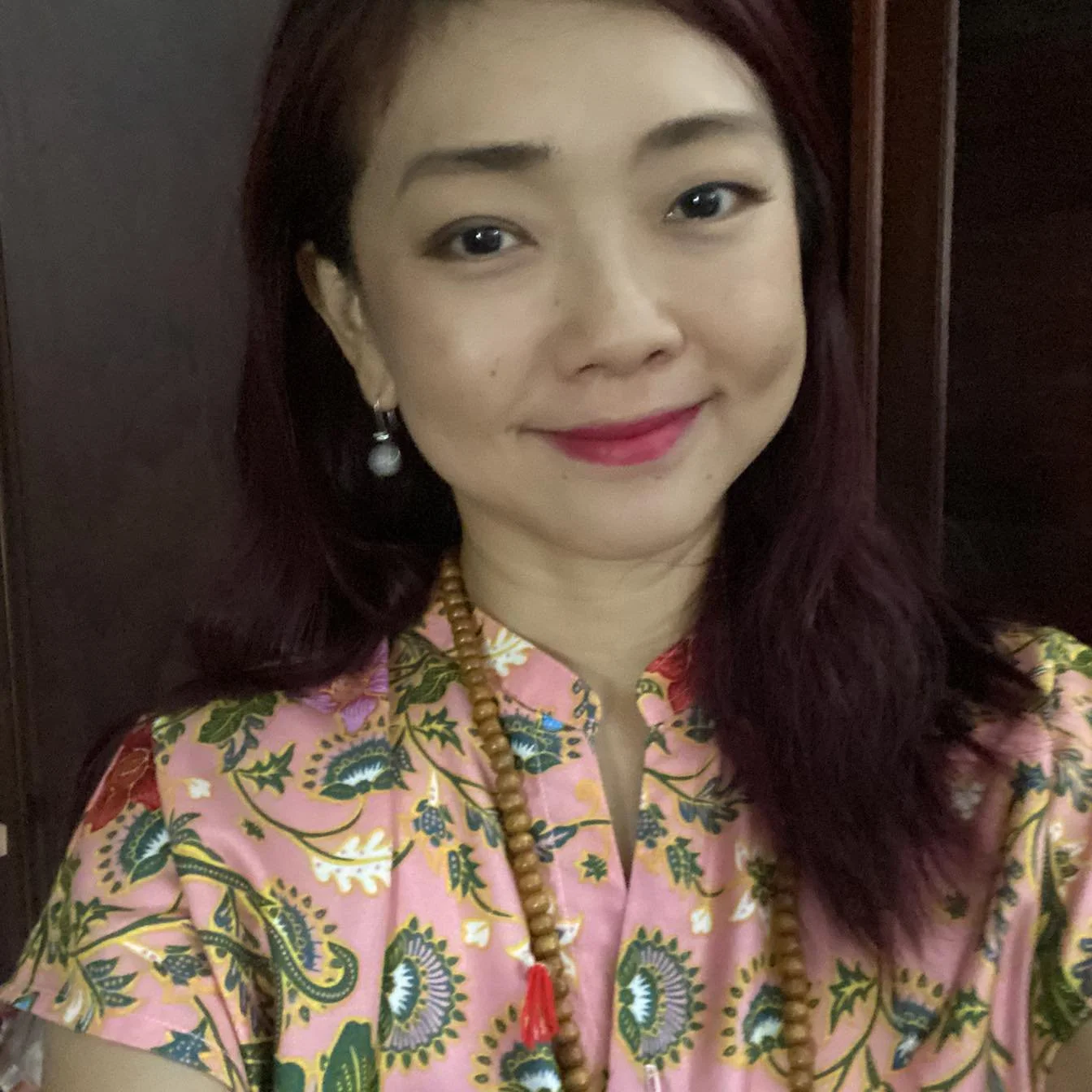 A woman with shoulder-length dark red hair, wearing earrings, a beaded necklace with a red tassel, and a pink floral-patterned top, smiling in front of a dark wooden background.