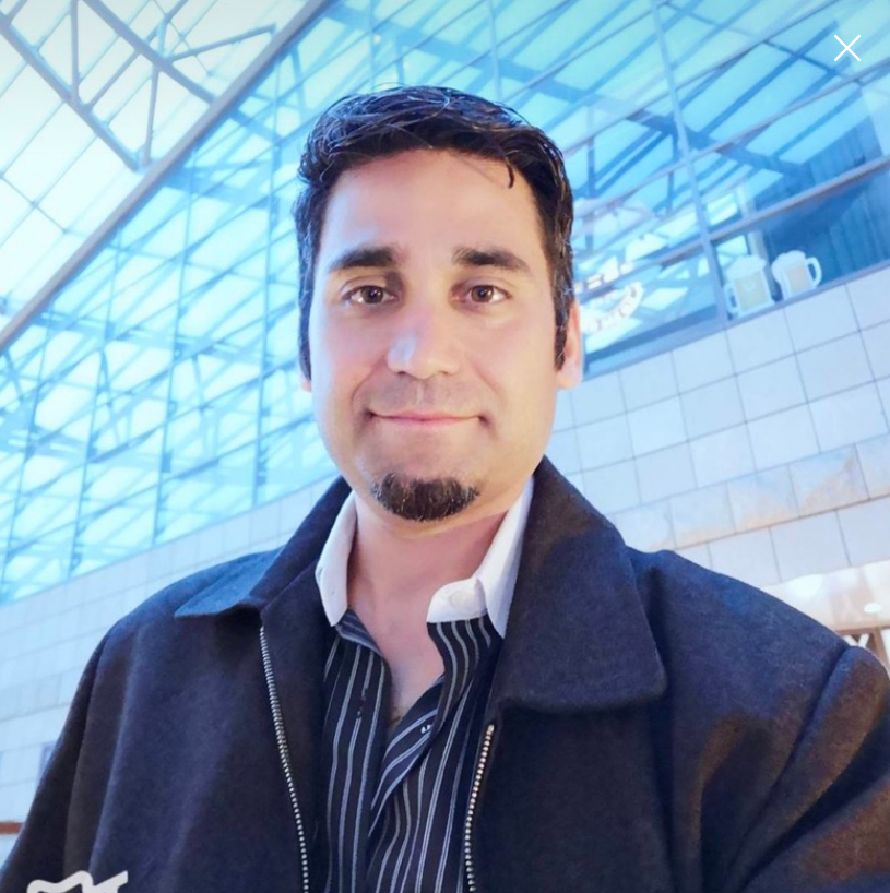 A man with dark hair and facial hair smiling at the camera indoors with a modern glass architecture ceiling in the background.
