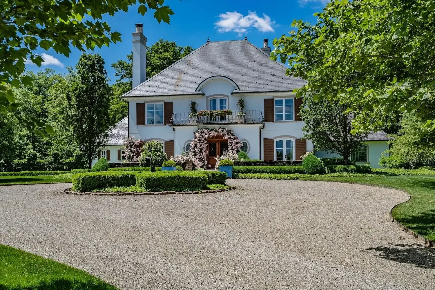 Elegant Tanglewood Homes white European-style country house with slate roof, wooden shutters, and balcony, framed by trees, hedges, flowering vines, and a circular gravel driveway.