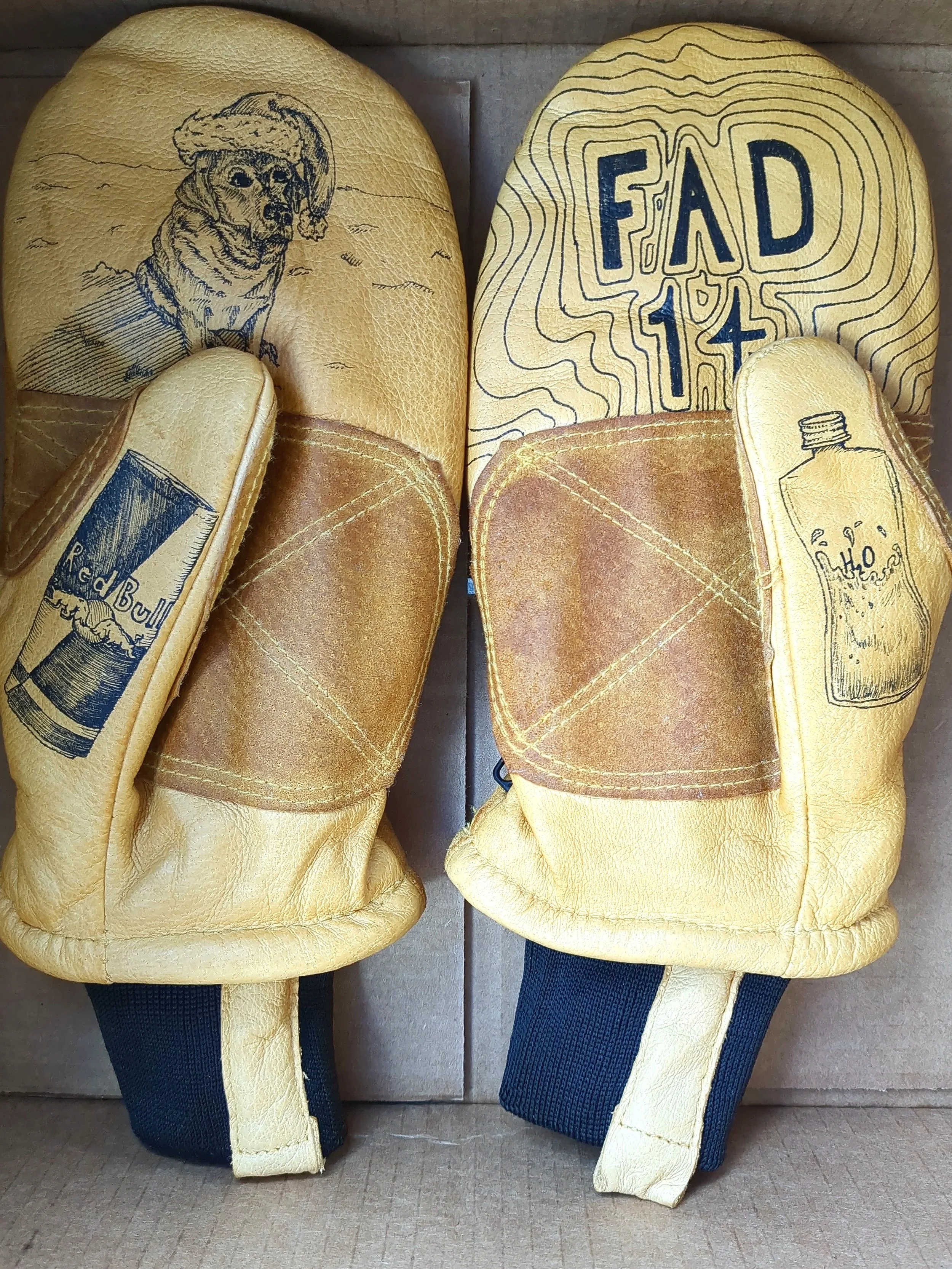 A pair of yellow leather gloves with printed designs of a dog wearing a hat, Red Bull energy drink can, a bottle of water, and the text 'FAD 14' on the back.