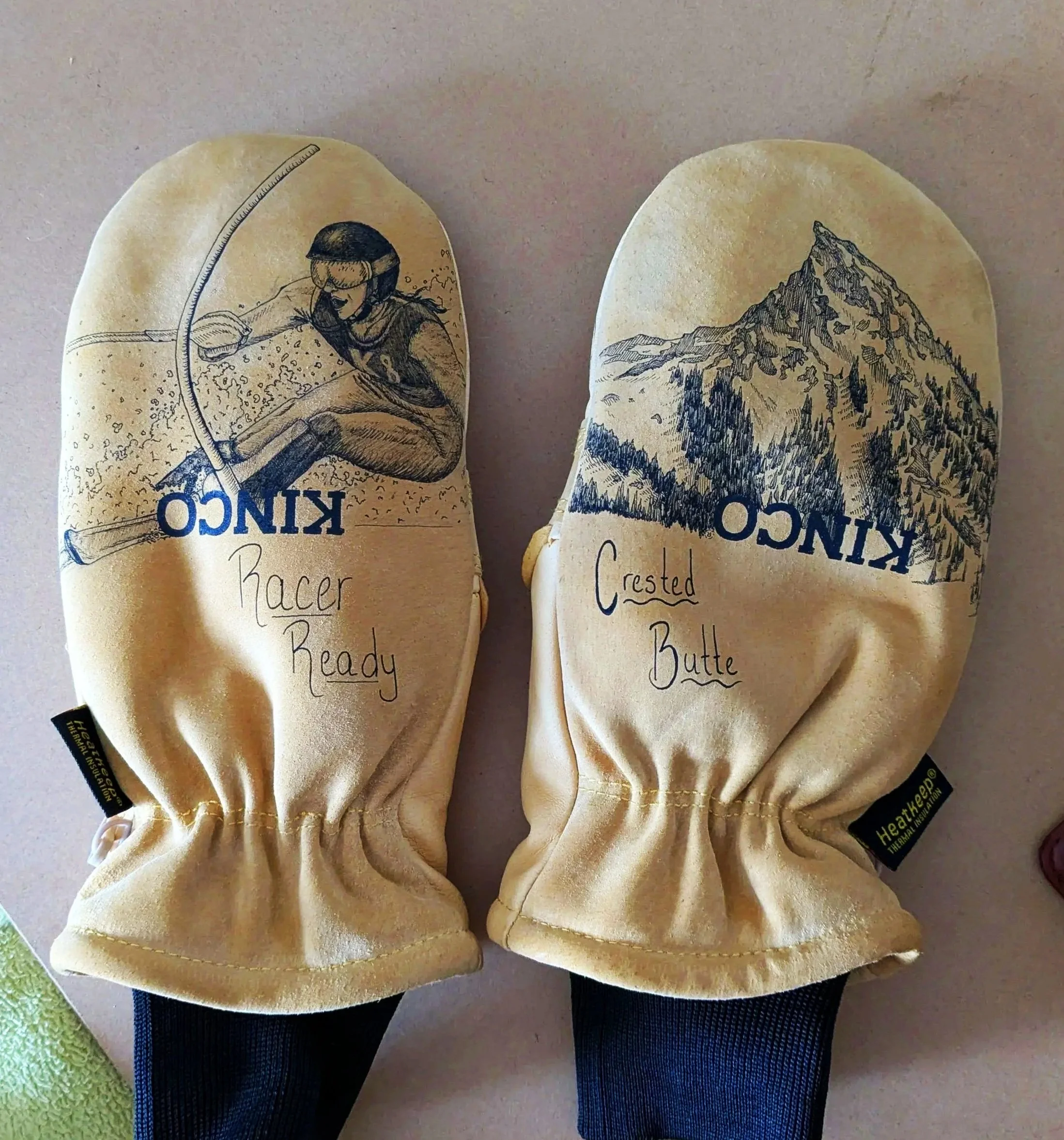 A pair of beige oven mitts with sketches and text. The left mitt has a skier illustration and says "Racer Ready." The right mitt has a mountain sketch and says "Crested Butte."