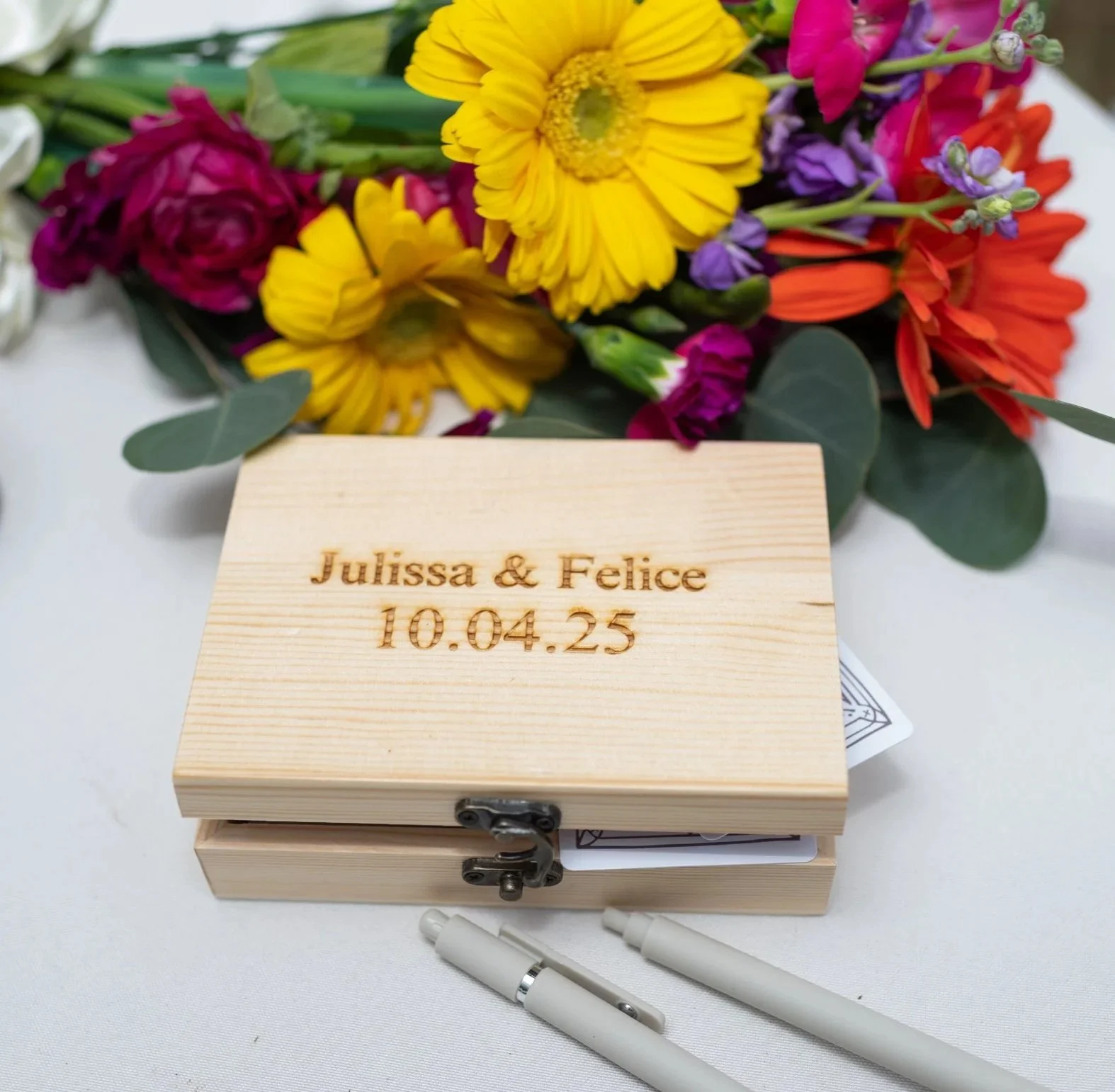 A wooden box with engraved text and a latch, with a colorful bouquet of flowers in the background.