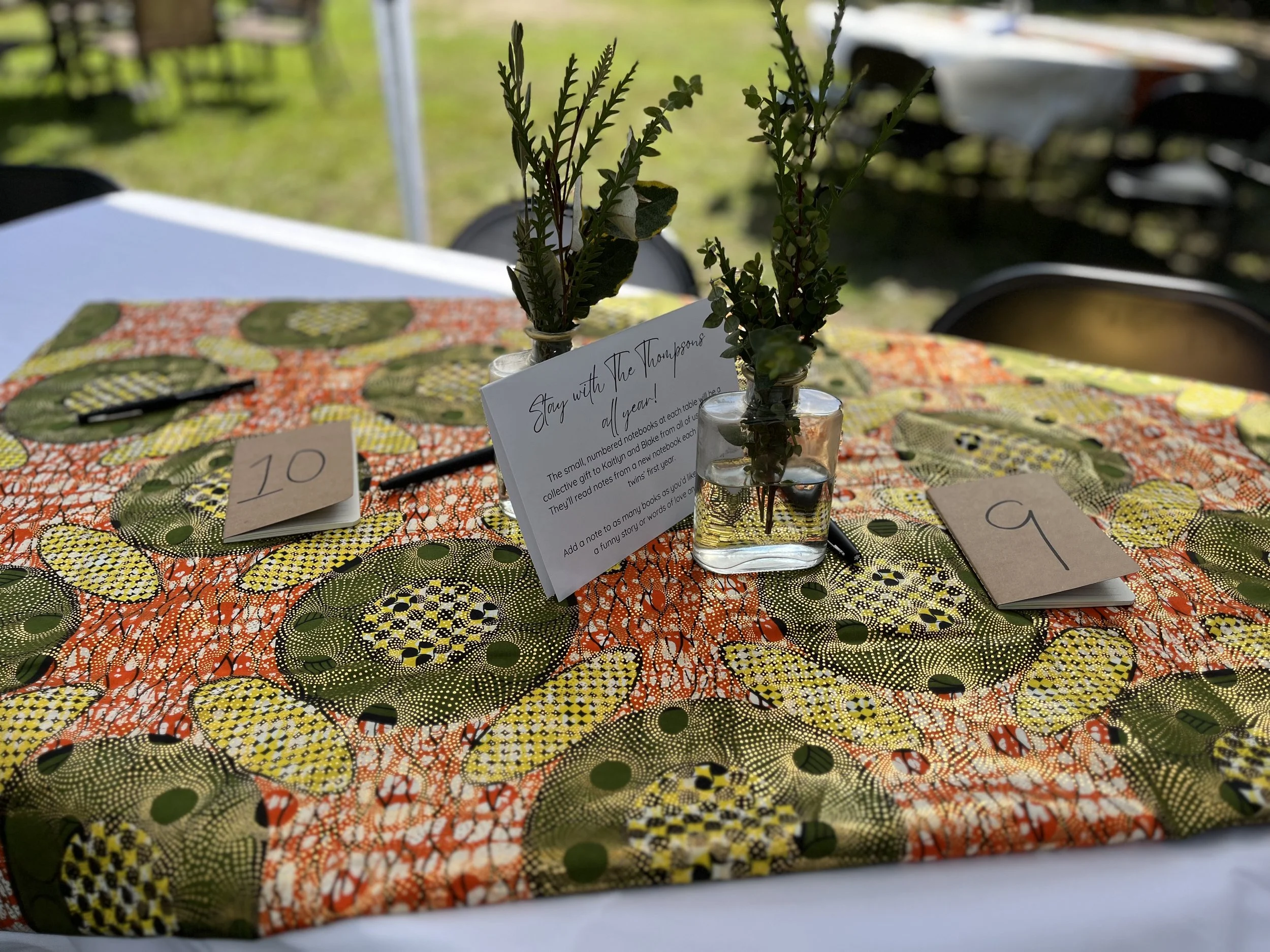 Vase with greenery on a table with a colorful patterned tablecloth, table number cards '10' and '9', and a note with handwritten text.
