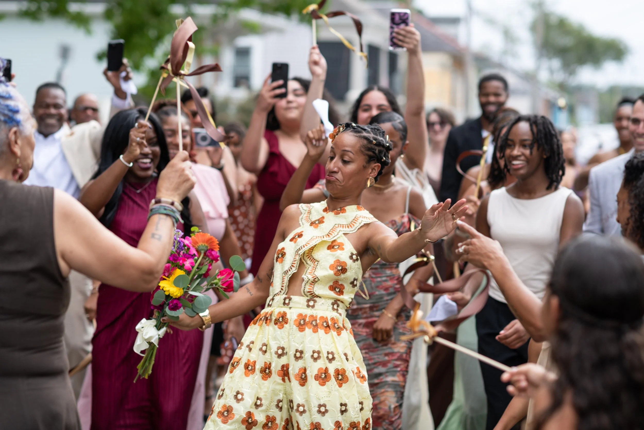 A woman in a floral dress holding a bouquet of flowers is dancing surrounded by a crowd of smiling people celebrating outdoors.