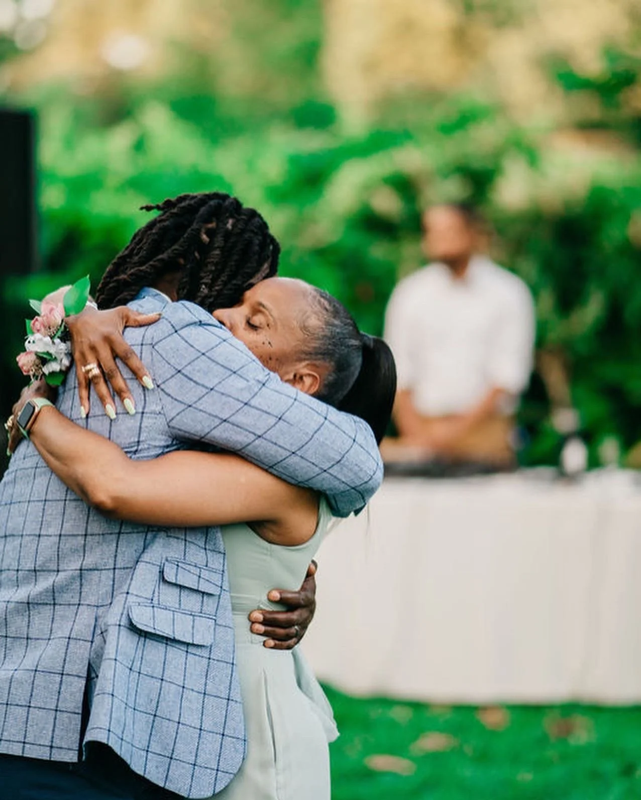 Two women are hugging each other tightly, expressing emotion, with lush green trees in the background. One woman is holding a small bouquet of flowers.