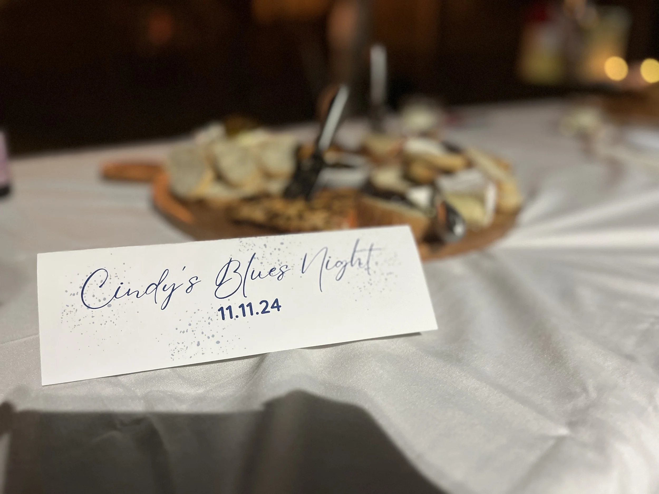 A place card with the words 'Cindy's Blues Night' and the date '11.11.24' written on it, placed on a white tablecloth with a cheese platter in the background.
