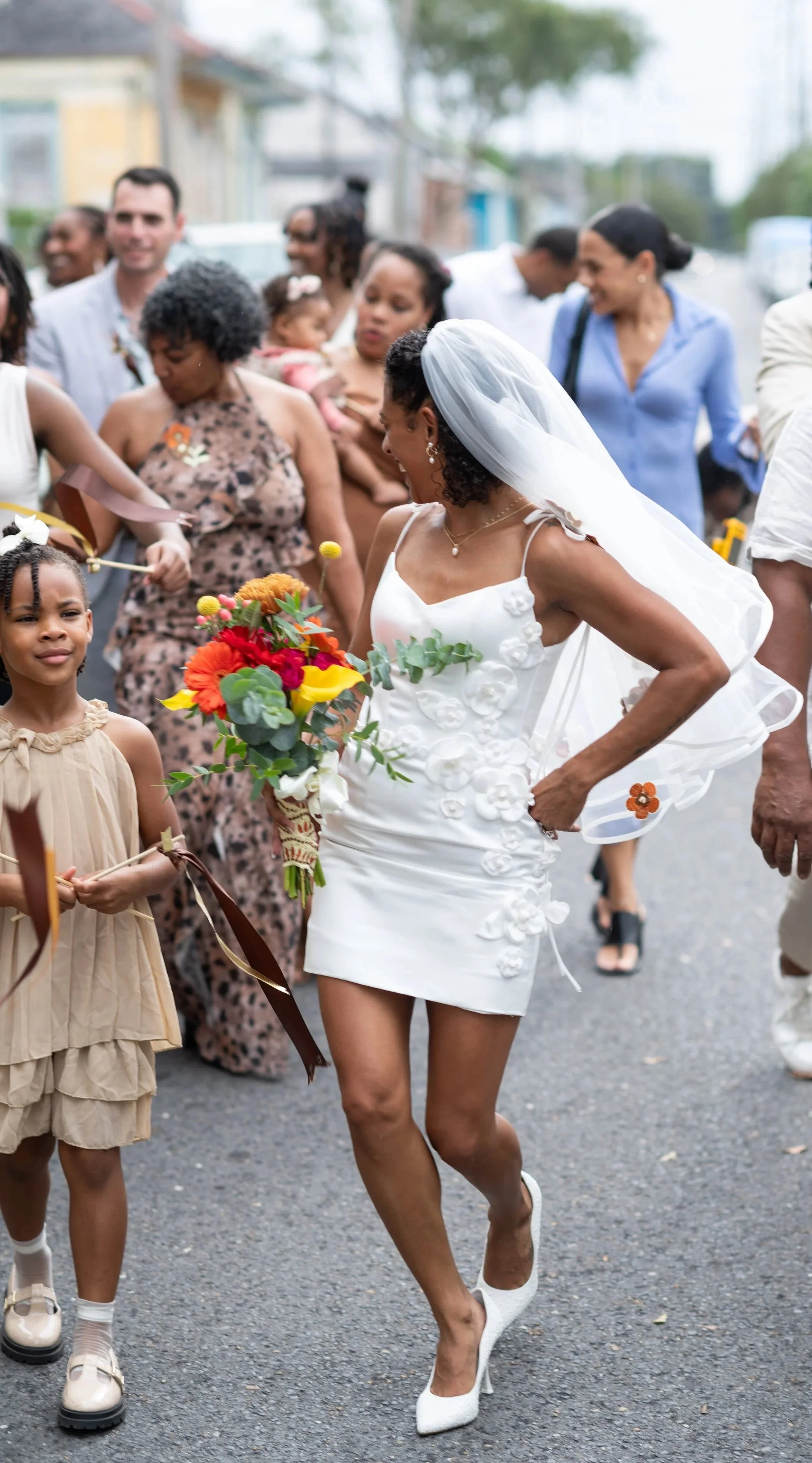 A bride in a white dress with floral details and a veil, holding a colorful bouquet, walking through a crowd of diverse people outdoors, some smiling and others looking on.