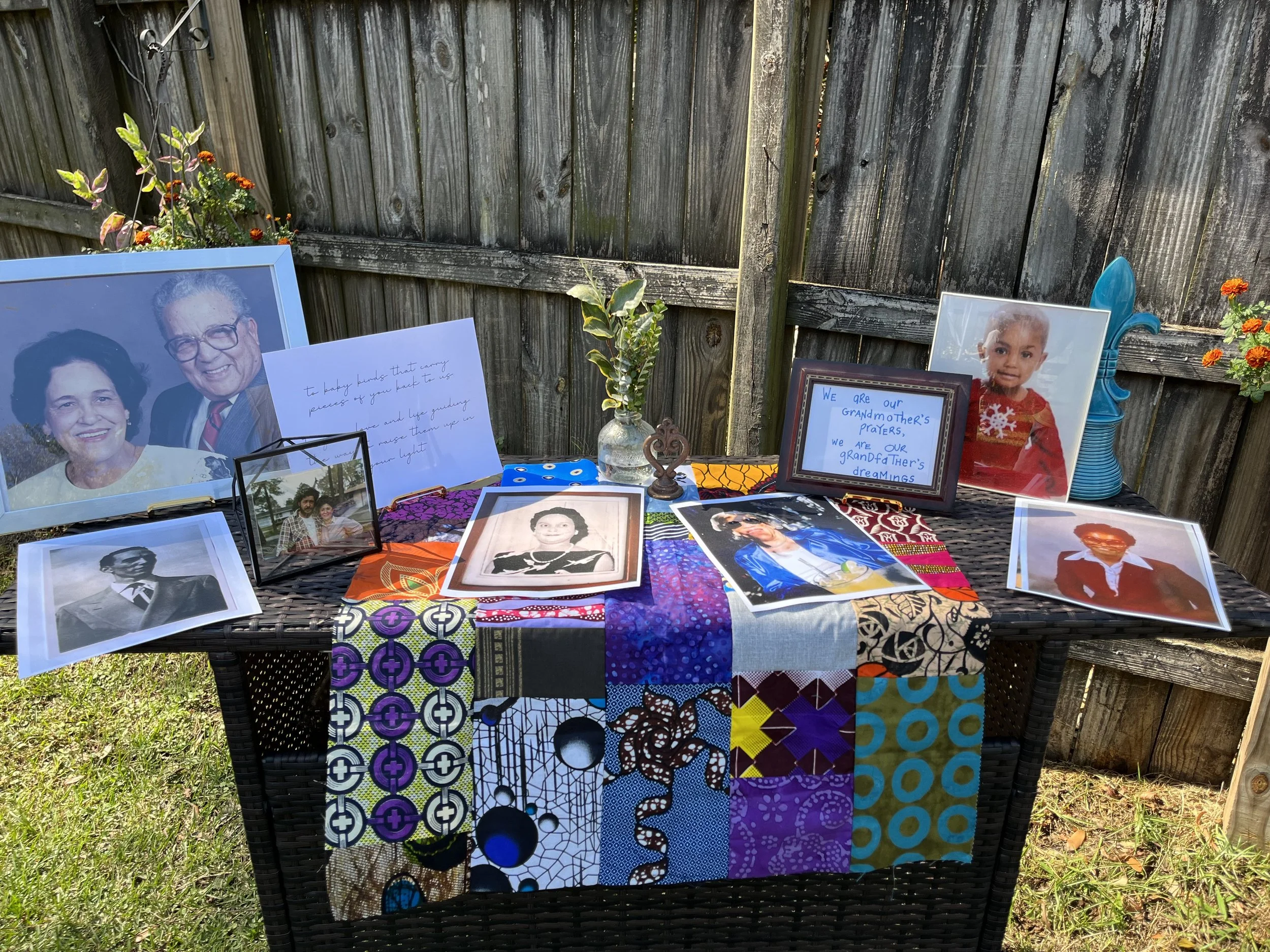 Memorial display with photographs, handwritten notes, and a framed message commemorating late family members, outdoors on a wicker table covered with a colorful quilt in front of a wooden fence and flowers.