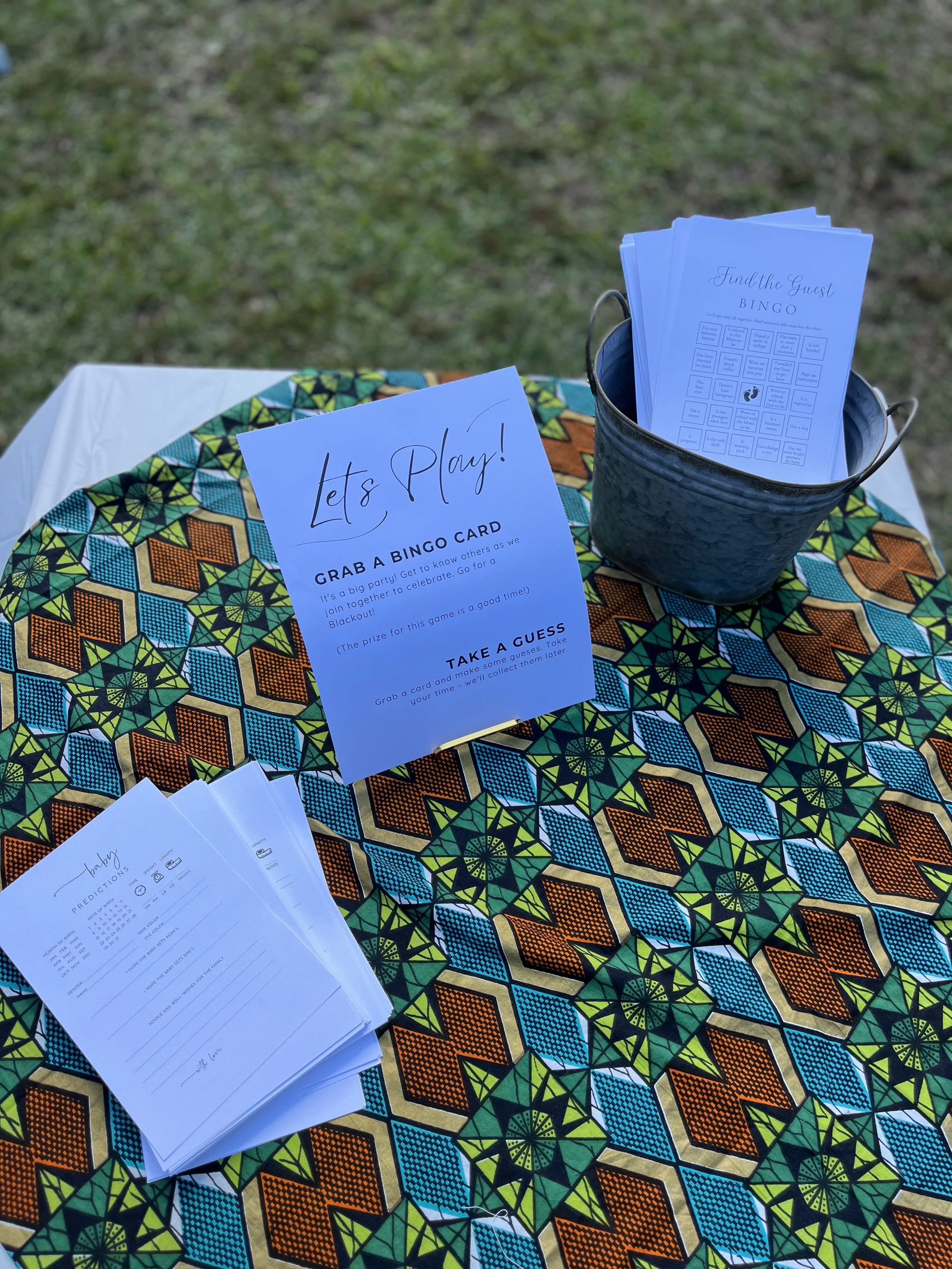 A table with a colorful geometric patterned tablecloth holds a sign, a stack of bingo cards, and a metal bucket with more bingo cards. The sign invites guests to play bingo and explains the game. The setting appears to be outdoors with grass visible 