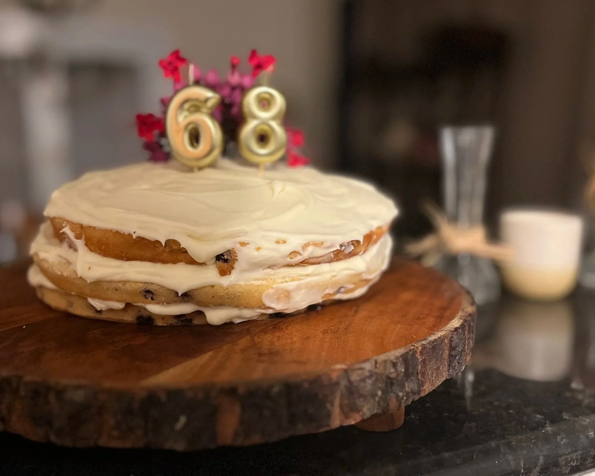 Layered birthday cake with white frosting and a gold and red candle topper with the number 68 on top, placed on a wooden cake stand with a blurred background.