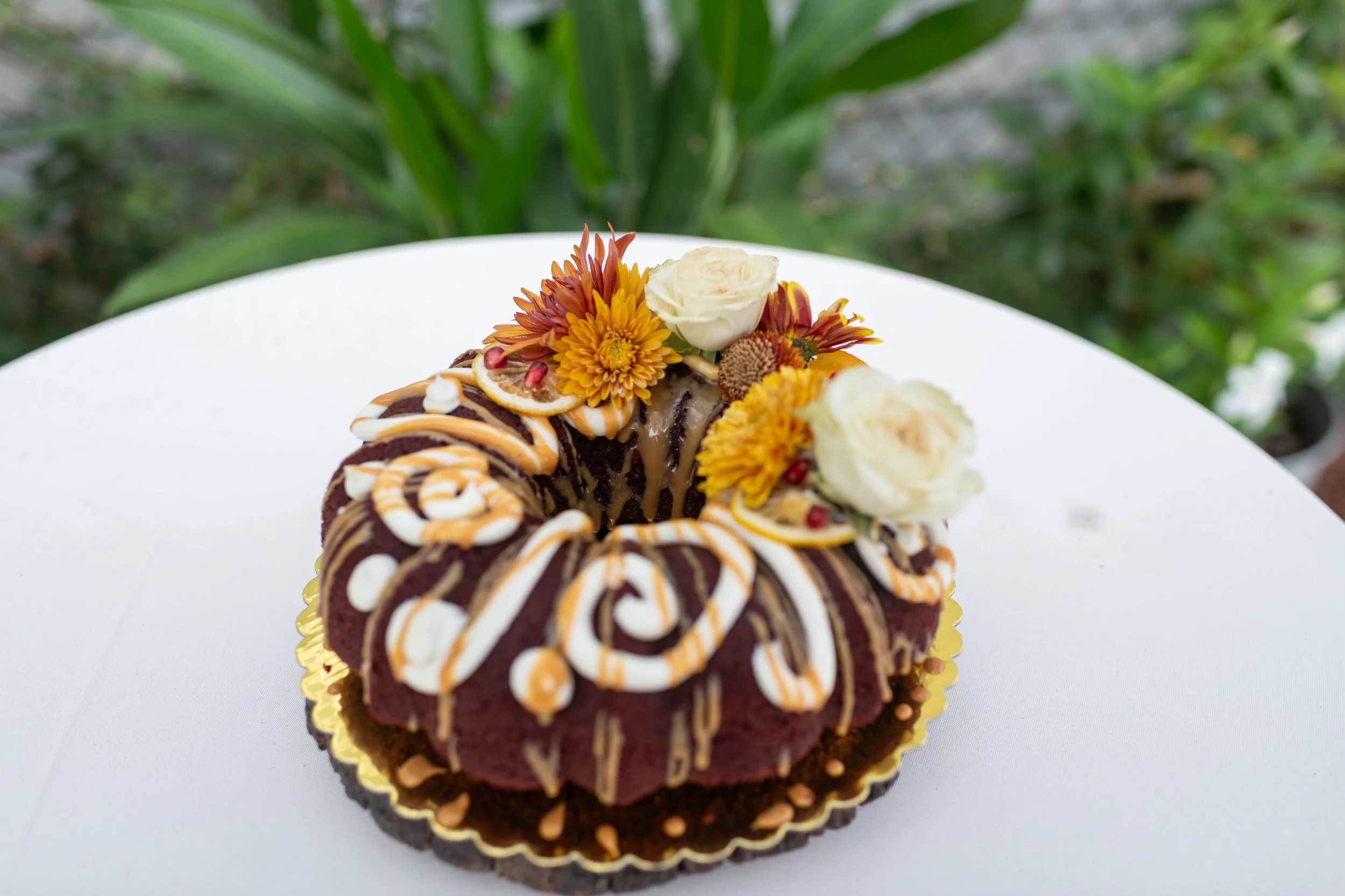 Chocolate bundt cake decorated with white and orange icing, and topped with white roses and yellow-orange flowers, on a white surface with a green garden background.