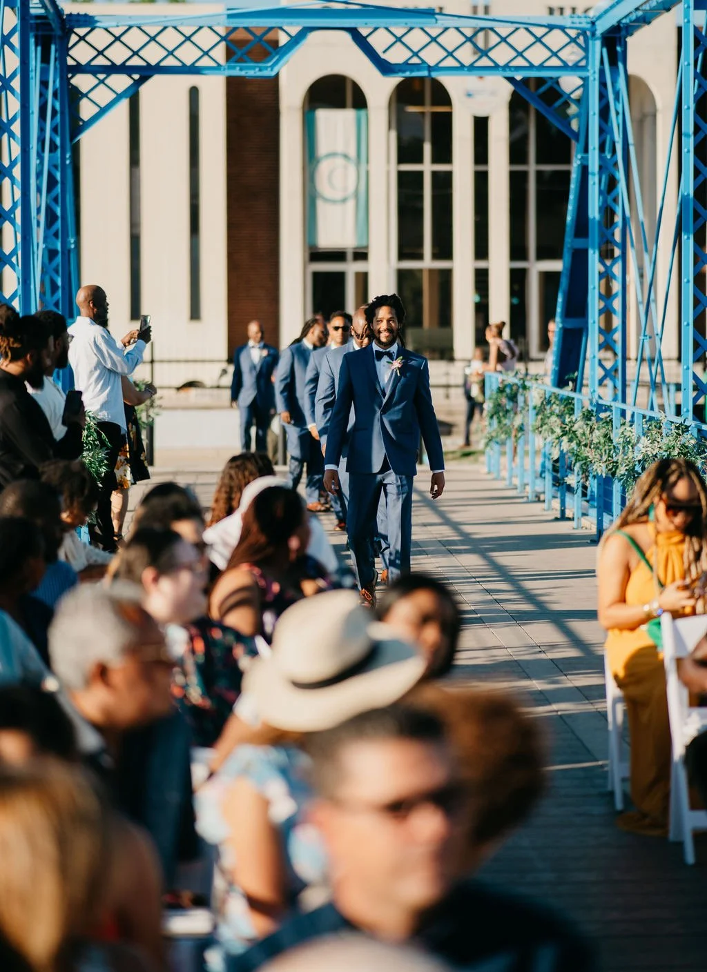 Wedding ceremony with a groom in a blue suit walking down the aisle, surrounded by guests, outdoors with a blue metal arch and a large building in the background.