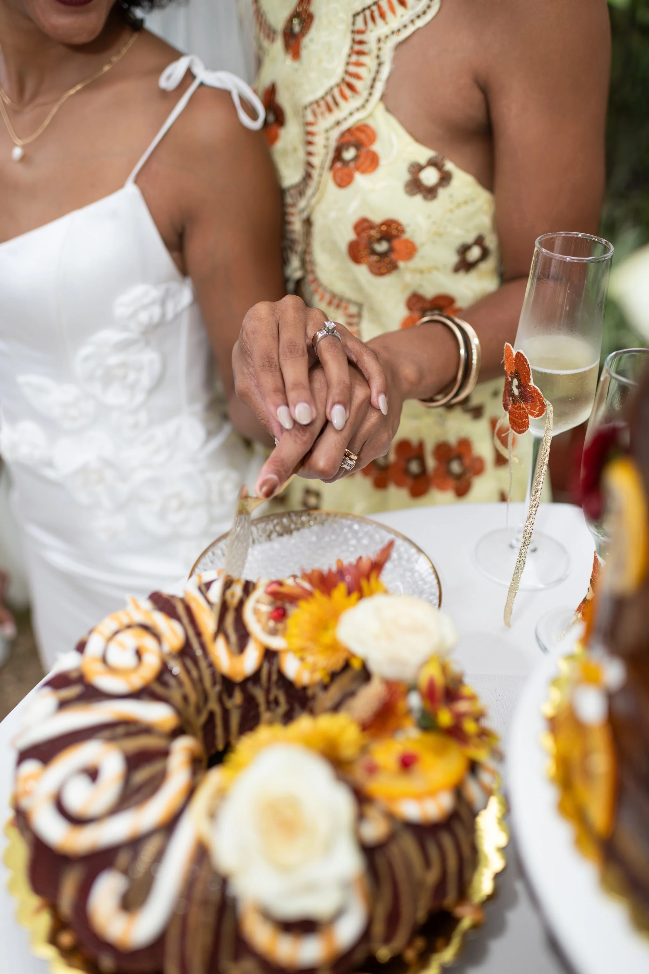 Two women holding hands during a celebration, with one woman wearing a white dress and the other wearing a yellow dress with orange and brown floral embroidery. They are cutting a decorated cake with flowers, alongside glasses of champagne on a table