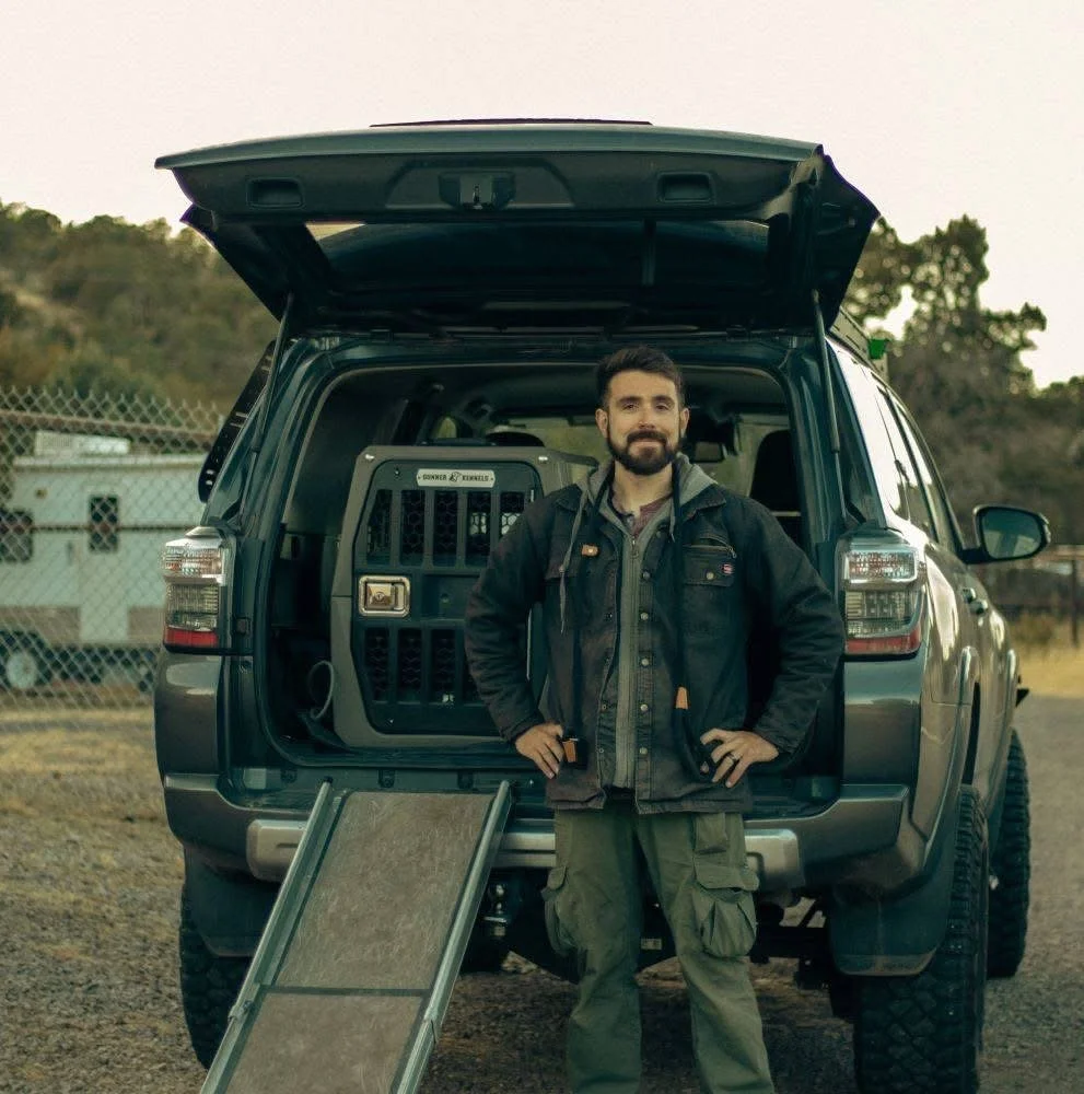 Man standing in front of an open SUV trunk with a pet cage inside, outdoors during daytime.