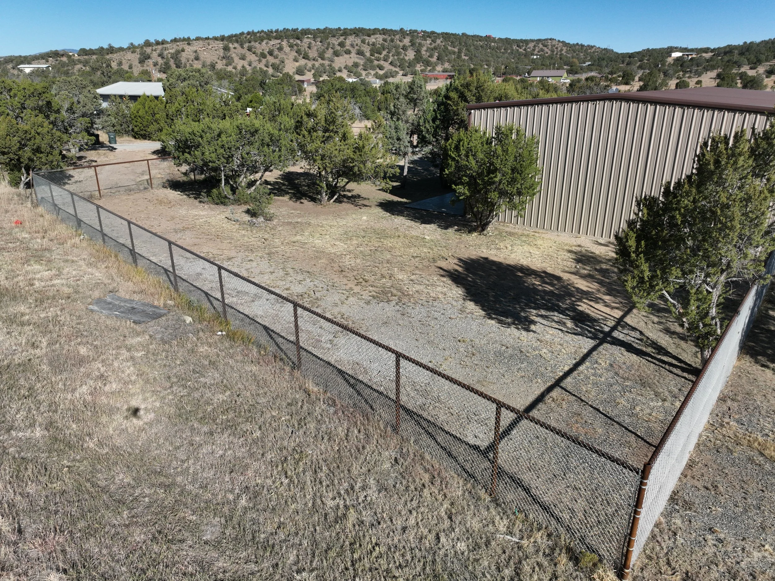 An empty fenced yard with trees, next to a large metal building, with hills and a clear blue sky in the background.