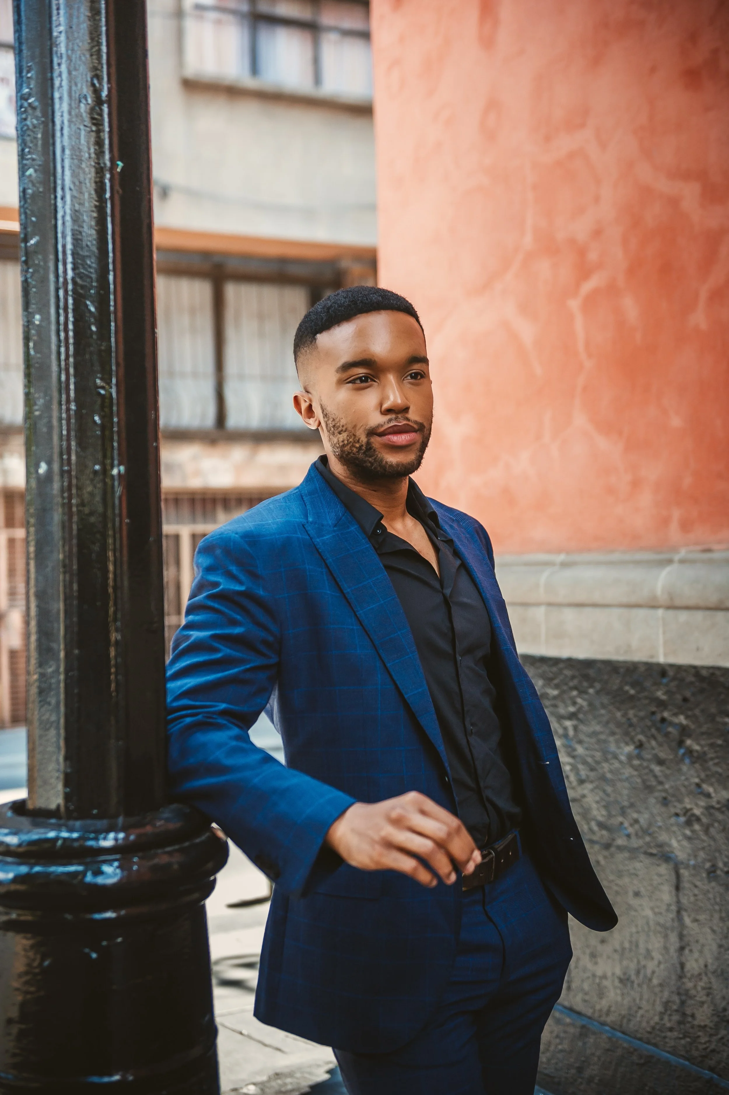A young man in a blue checkered suit and black shirt standing near a black pole on a city street, with a building with large windows and a peach-colored wall in the background.
