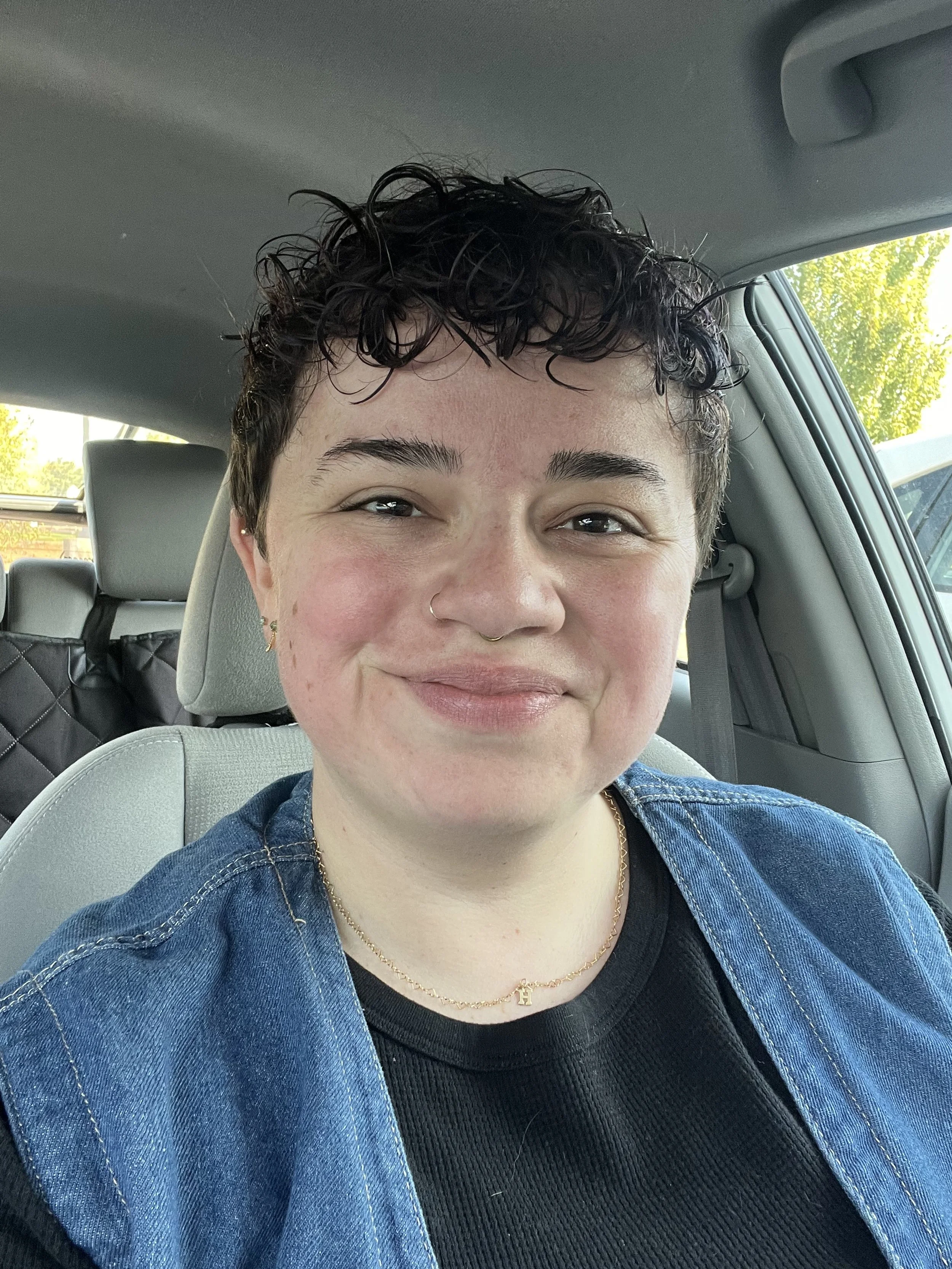 A person with short curly hair and a nose ring, smiling inside a car, wearing a black shirt, a blue denim jacket, and a gold necklace.
