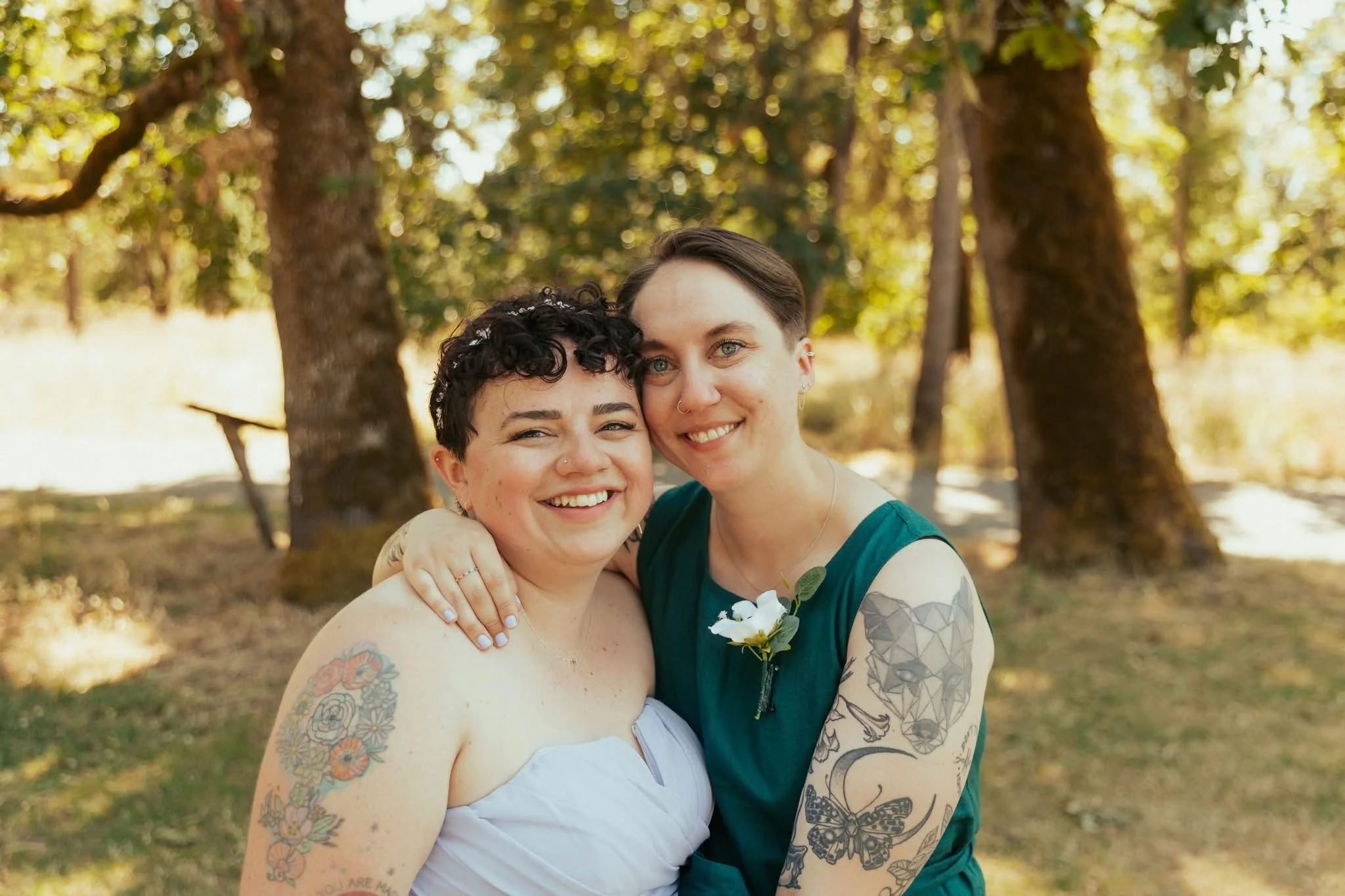 Two women smiling and hugging outdoors among trees, one in a white dress and the other in a dark green dress, both with tattoos and short hair, with sunlight filtering through the leaves.