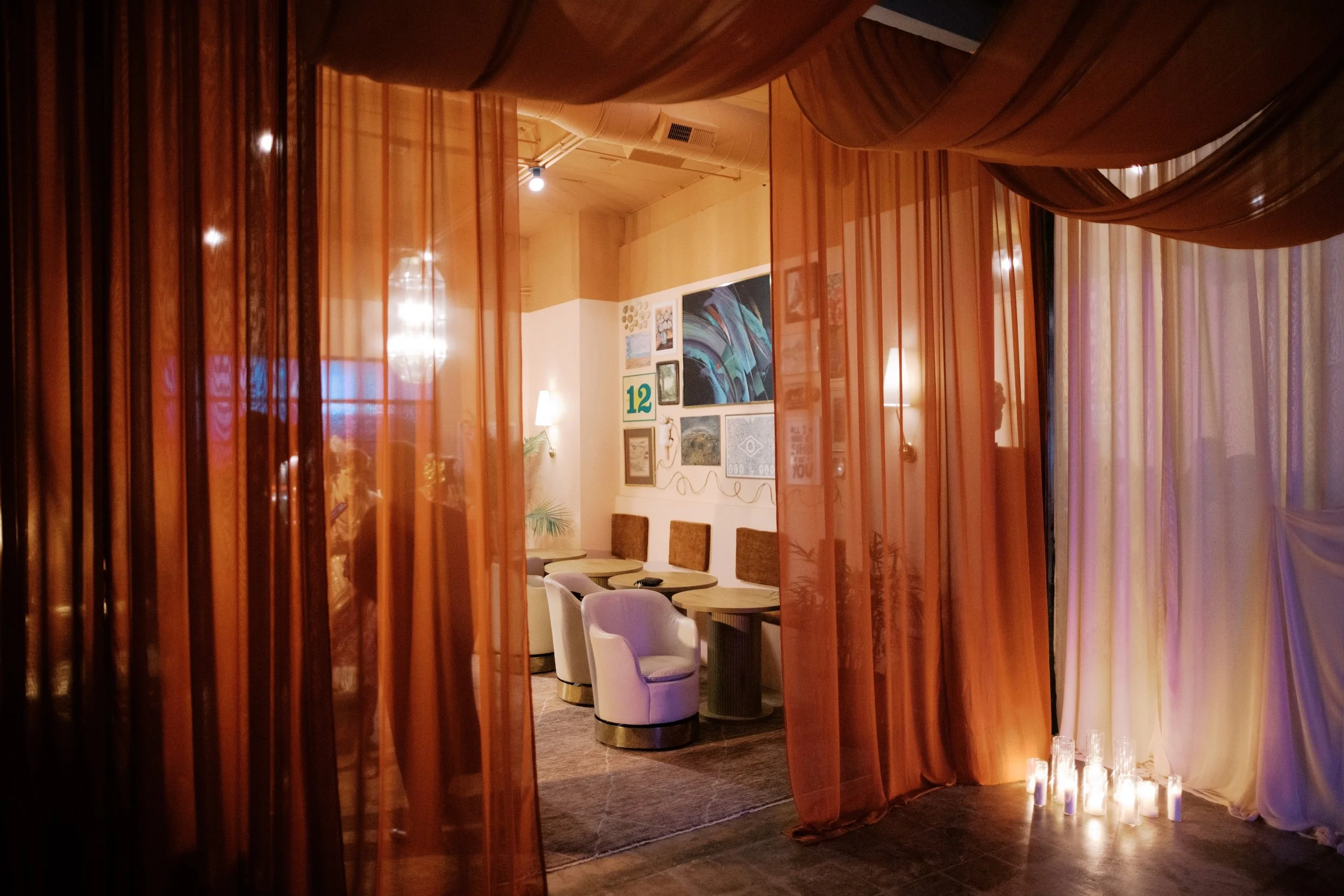 A cozy room with orange curtains, white chairs, small tables, wall art including paintings and a green "12" sign, and candles on the floor, creating a warm ambiance.