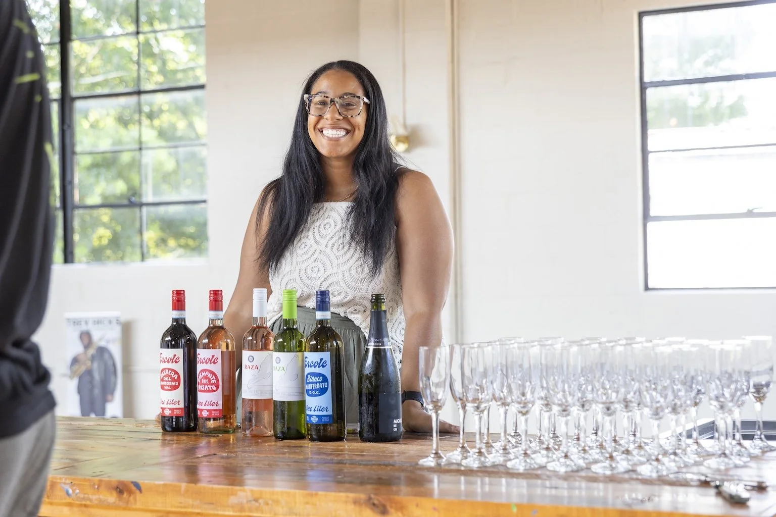 Woman with long dark hair, glasses, and a white lace dress smiling behind a table with bottles of wine and empty champagne glasses in a bright room.
