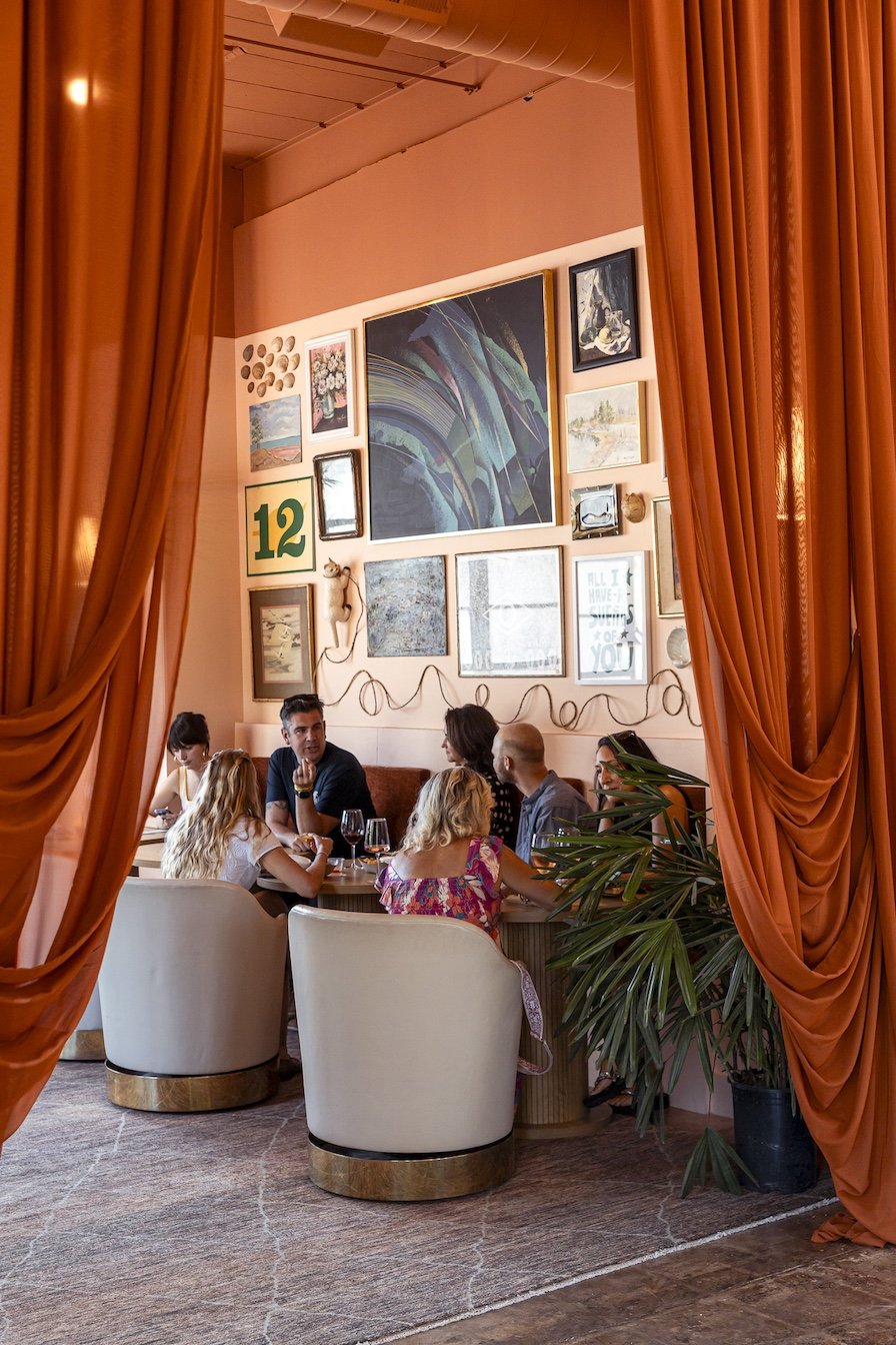 People sitting at a dining table in a restaurant with orange curtains, pink walls, and gallery wall art.