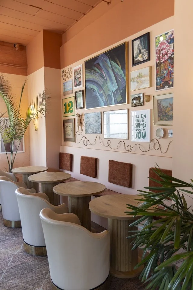 Interior of a cozy cafe or lounge with a wall decorated by various framed artworks and a green number 12 sign. There are five white chairs with rounded backs and wooden tables arranged along the wall. Large green plants are in the foreground and to t