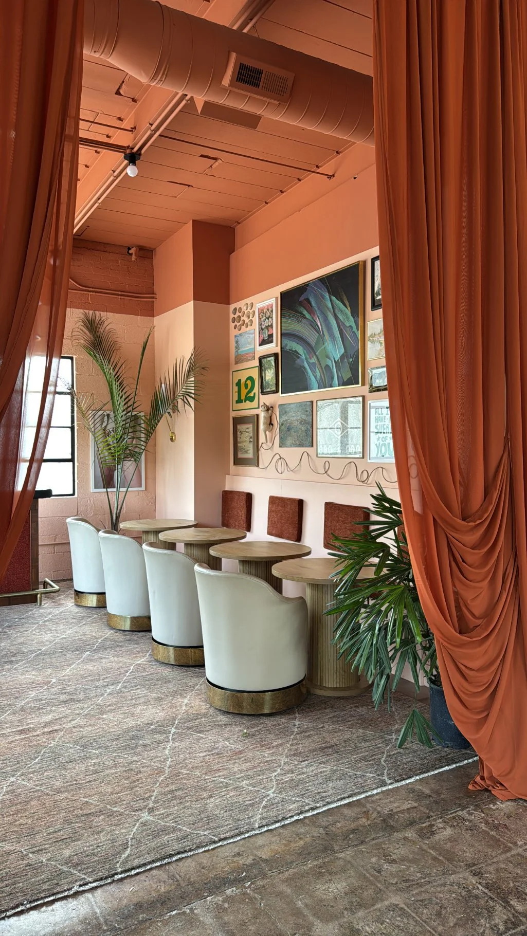 Lounge area with white chairs around a curved wooden table, a large potted plant, coral-colored curtains, and a gallery wall with various framed pictures and artwork.