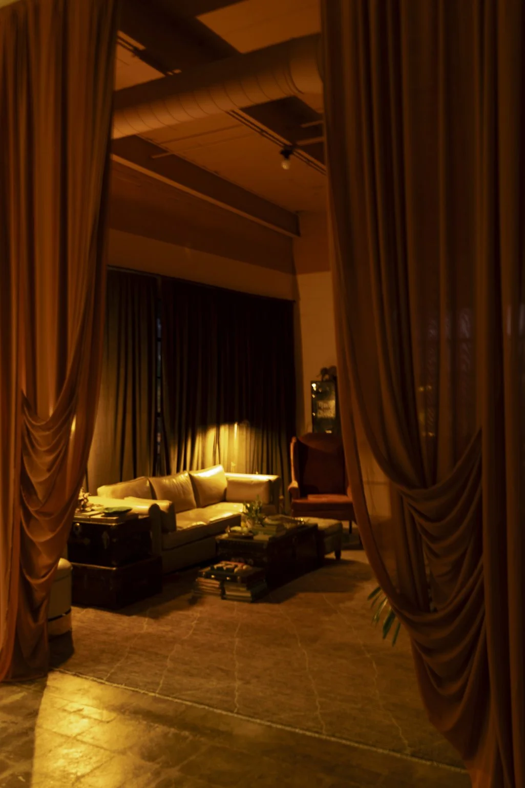 A cozy living room with a beige sofa, a coffee table with books, and dark curtains, viewed through open curtains with warm lighting.