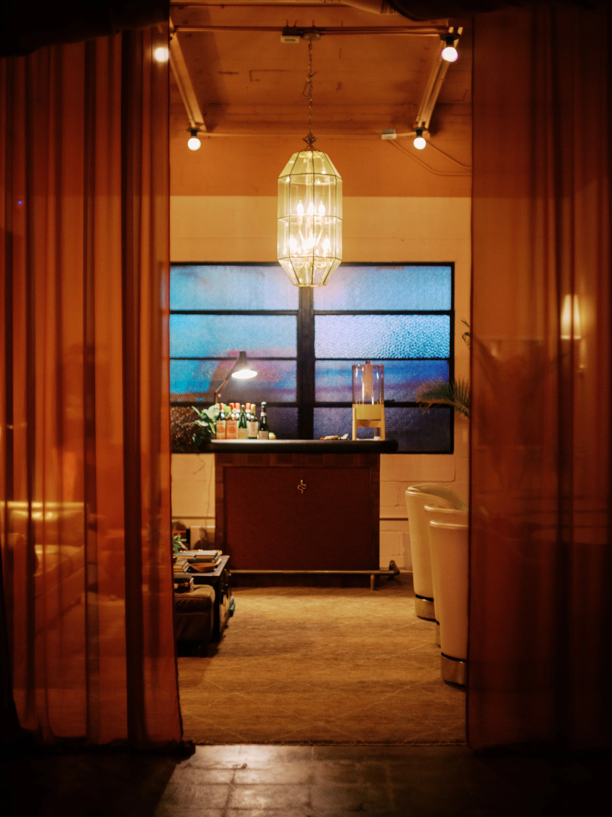 View into a cozy, dimly lit room with a hanging chandelier, bar area with bottles, a glass dispenser, and a window with frosted glass panes.