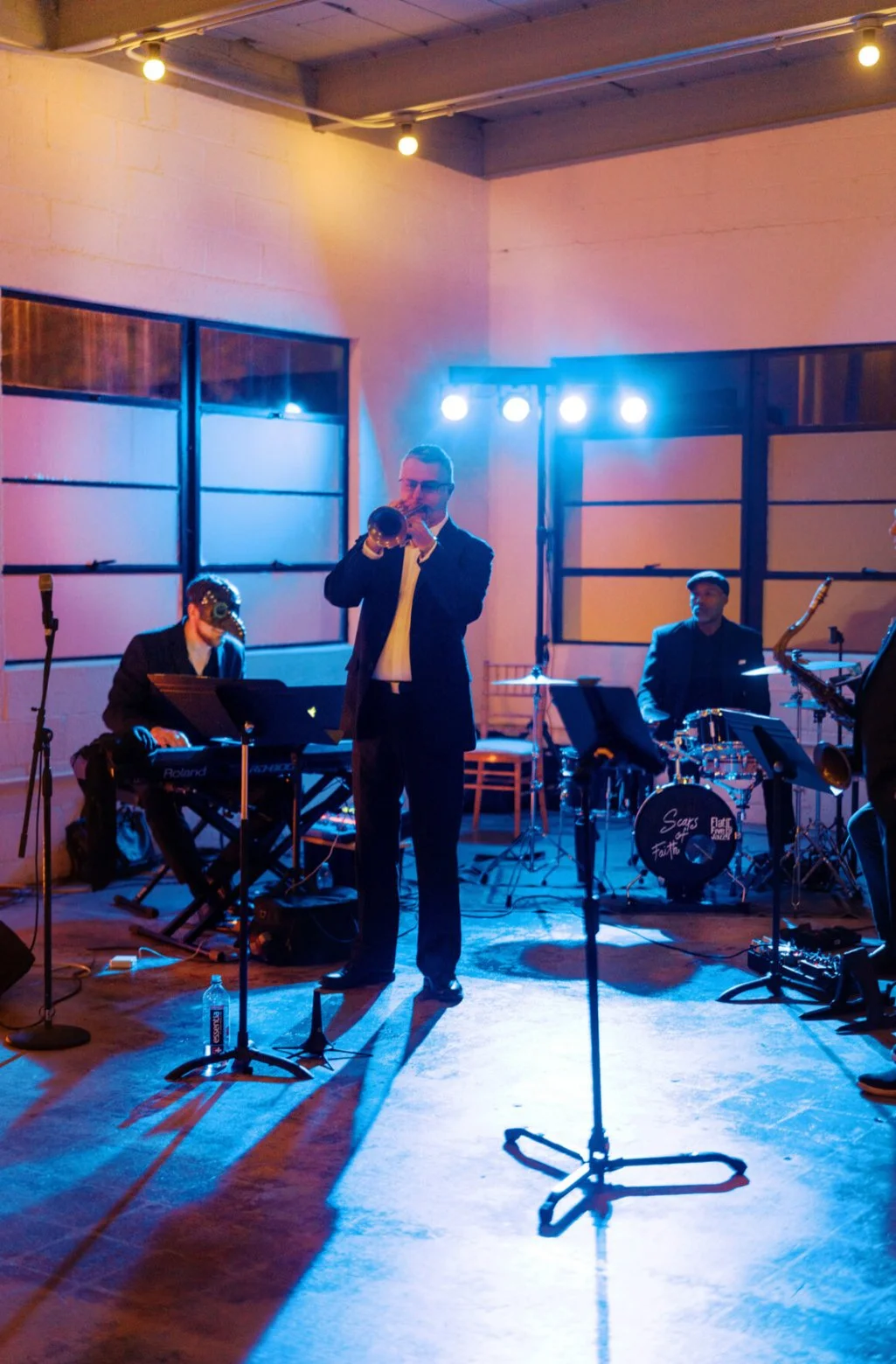 A jazz band performing on stage with a singer playing the trumpet, a keyboardist, and a drummer under colorful stage lights.