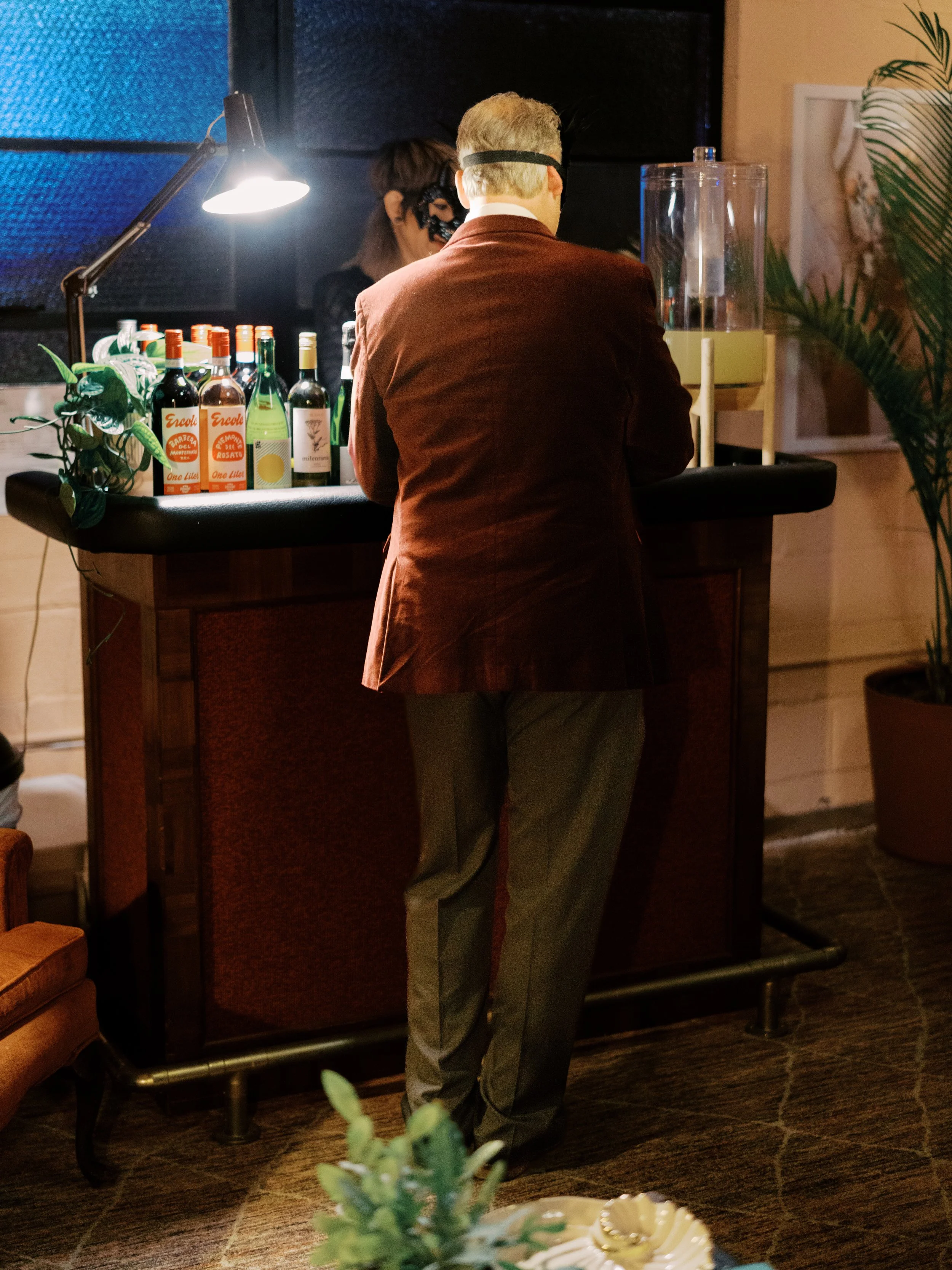 A person with blonde hair, wearing a brown blazer, stands at a bar with bottles of alcohol, a lamp, and a drink dispenser. In the background, a person with dark hair and a face mask is behind the bar.