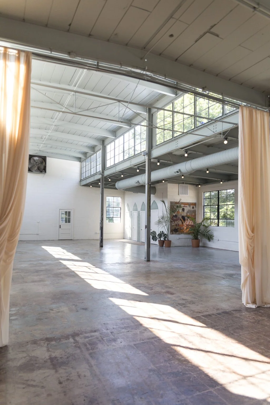 Casa Nueva , an industrial-style event space with high ceiling, large windows, potted plants, and artwork on wall, illuminated by natural light.